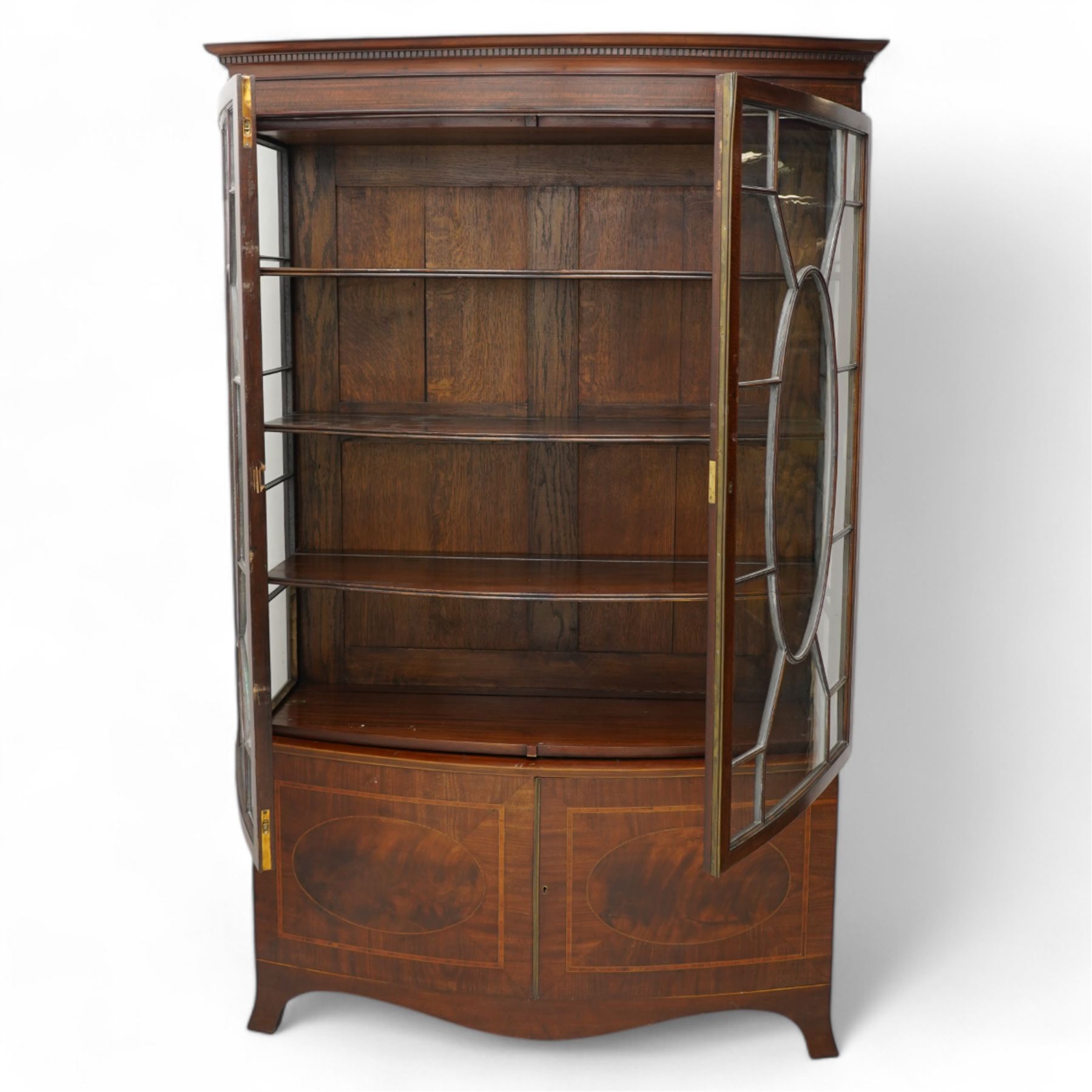 Edwardian mahogany and satinwood bow-fronted display cabinet, moulded dentil cornice over two astragal glazed doors, fitted with three shelves, double cupboard below, the doors inlaid with figured oval panels and satinwood bands, shaped apron on splayed bracket feet 
