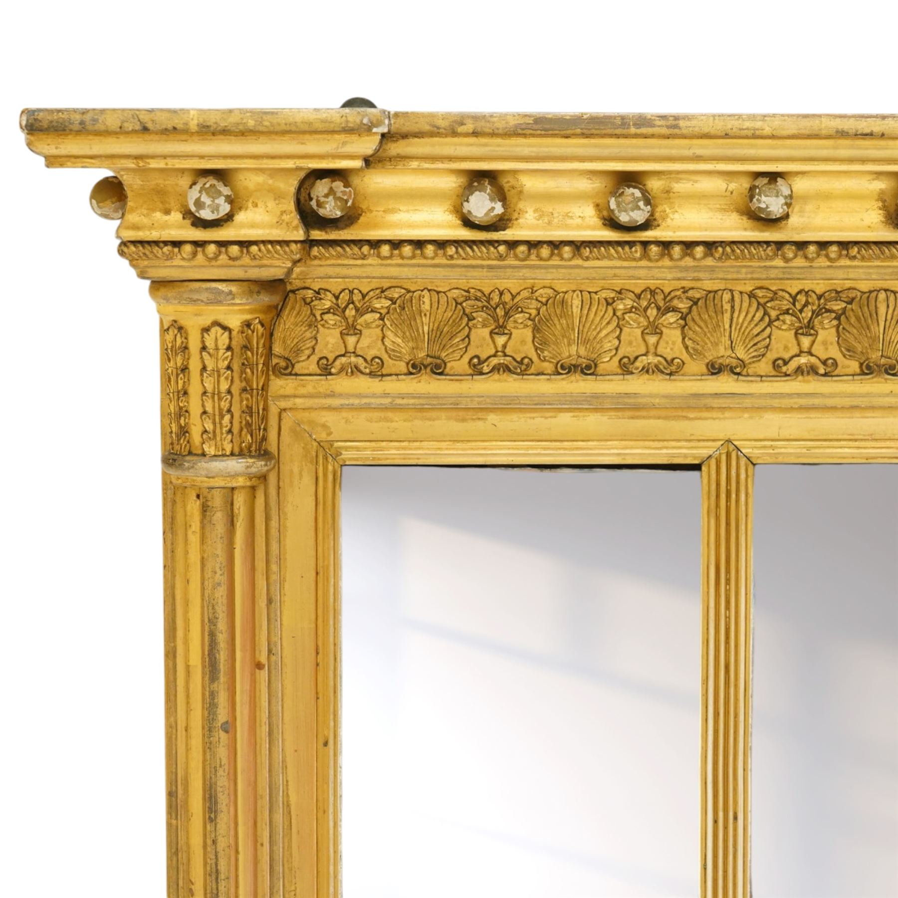 Regency period gilt and gesso framed tripartite overmantel mirror, projecting cavetto cornice applied with globular mounts, the frieze carved with repeating scallop and floral urn motifs, flanked by canted cluster-column pilasters with acanthi capitals, the triple rectangular bevelled plates within reeded slips, on skirted ebonised base
