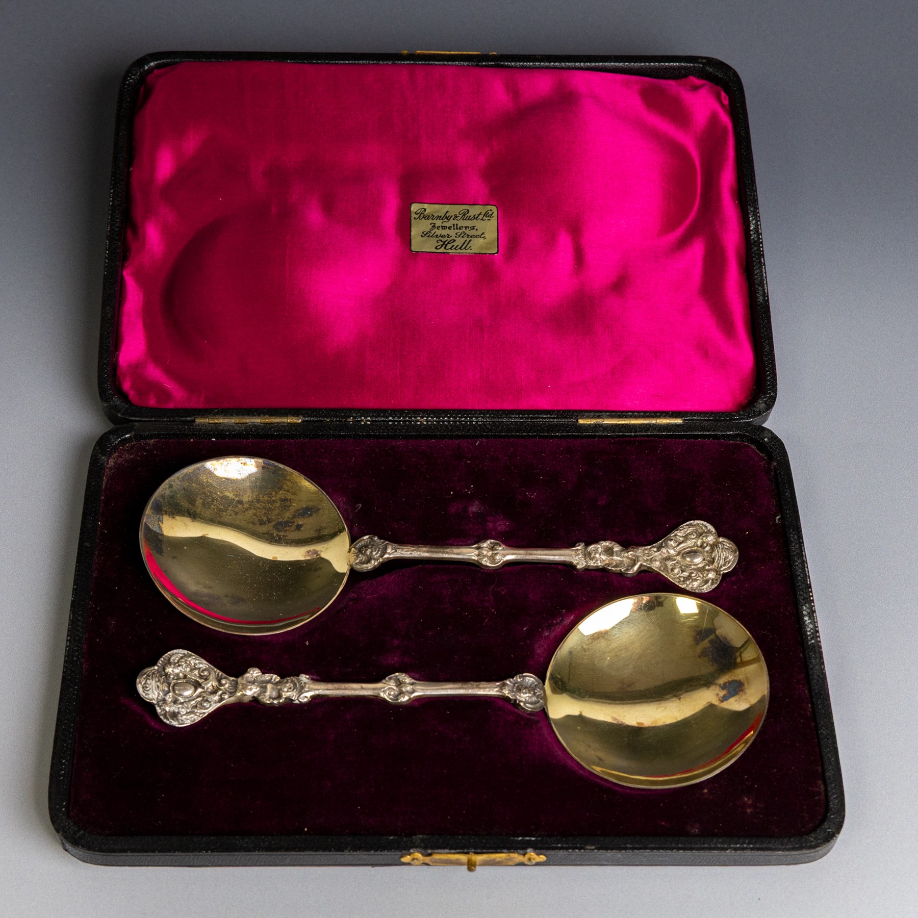 Pair of late Victorian silver serving spoons, each with circular gilt bowls and embossed putti decoration, hallmarked William Hutton & Sons Ltd, London 1895, contained within a tooled leather box with burgundy velvet and silk interior