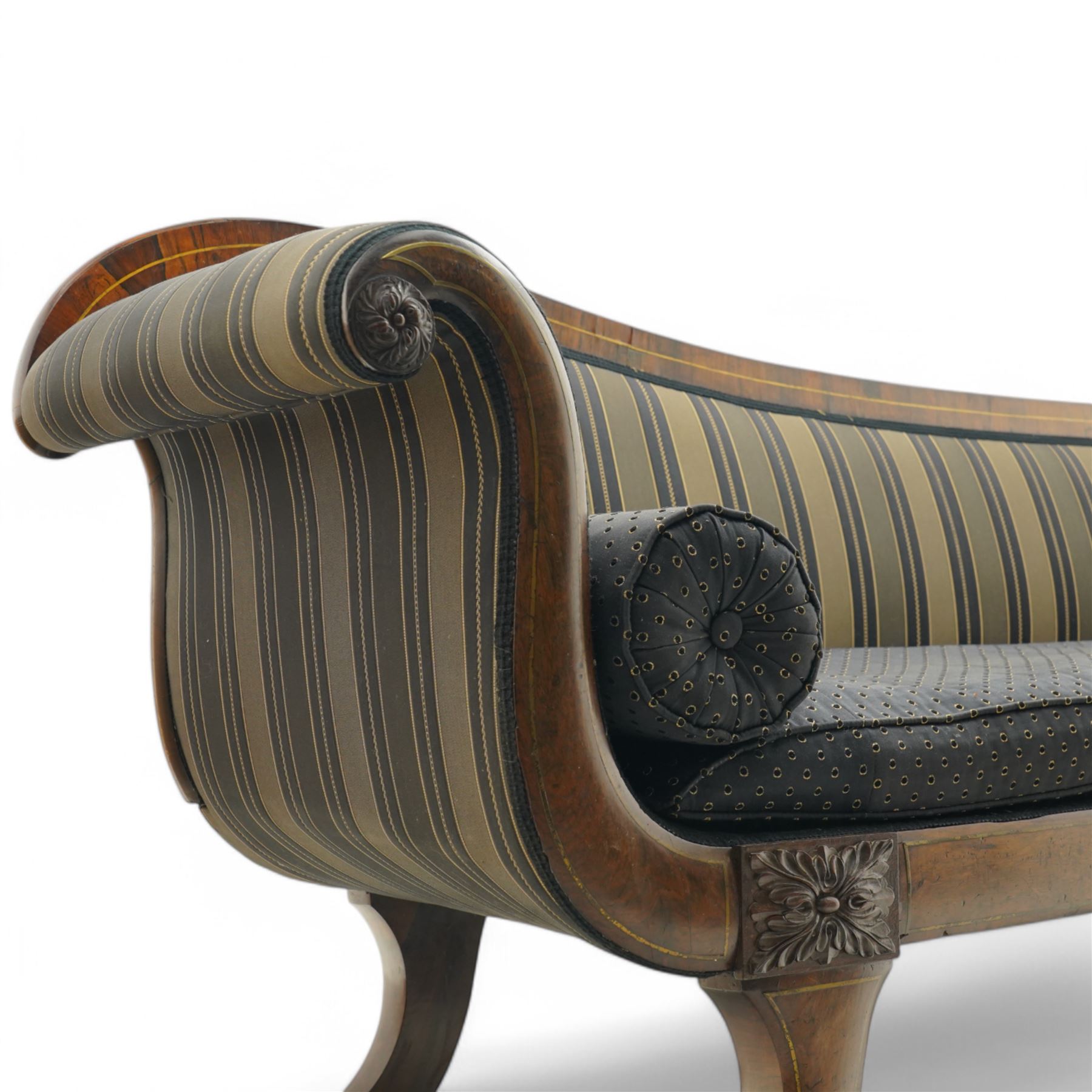 Regency brass inlaid rosewood chaise lounge or settee, shaped back over rolled head and foot, upholstered in black and gold striped fabric with rectangular squab cushion and bolster cushion, the rolled terminals carved with acanthus leaf roundels, lower rectangular foliate carved panels, on out splayed supports with paw carved feet 