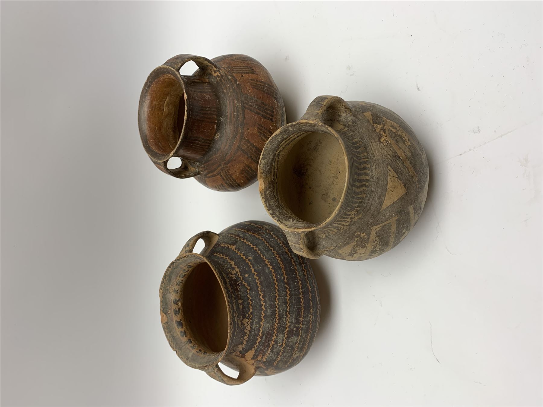 Three Chinese Neolithic pottery vessels, each with twin loop handles to the shoulder and painted geometric banded decoration to the body, all bearing labels for Eskenazi London, tallest H14cm