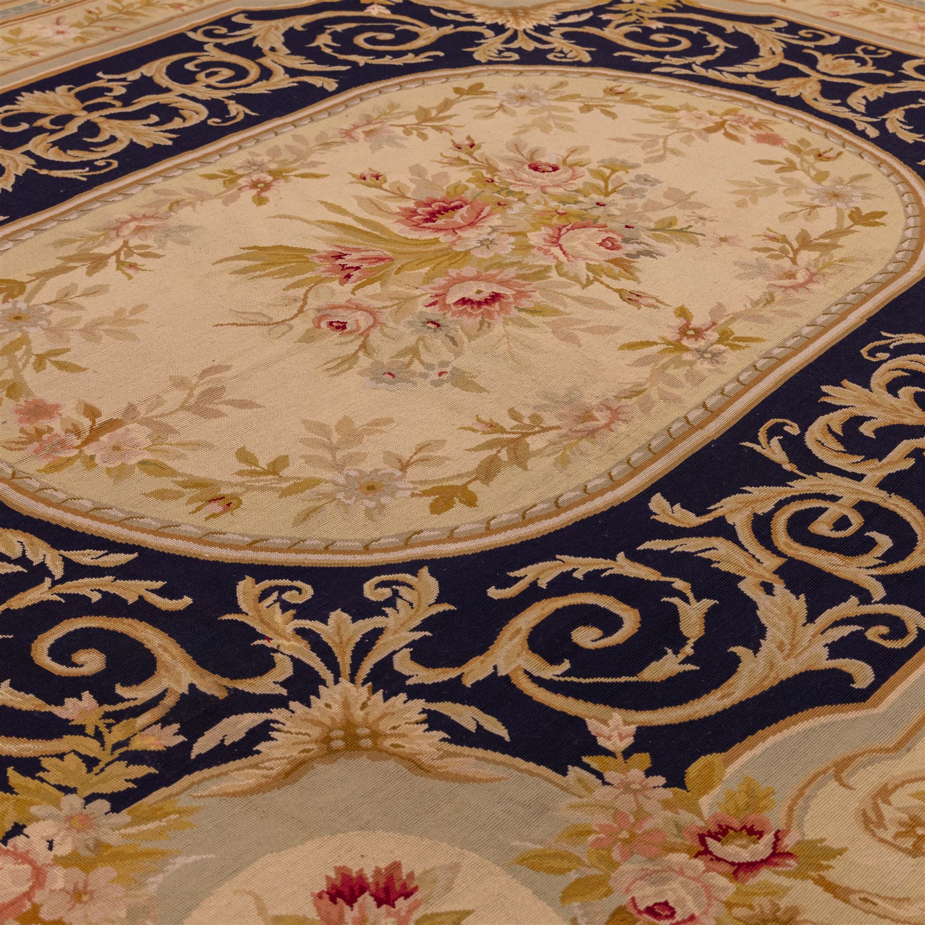 20th-century Aubusson-style woven tapestry wall hanging of rectangular form, the design with a central oval floral medallion composed of delicate sprays in soft pinks, creams, and muted green enclosed by an elaborate dark blue scrolling acanthus ground enriched with foliate ornamentation, field framed by floral cartouches to the corners and multiple guard borders in pale blue, ivory, and ochre tones