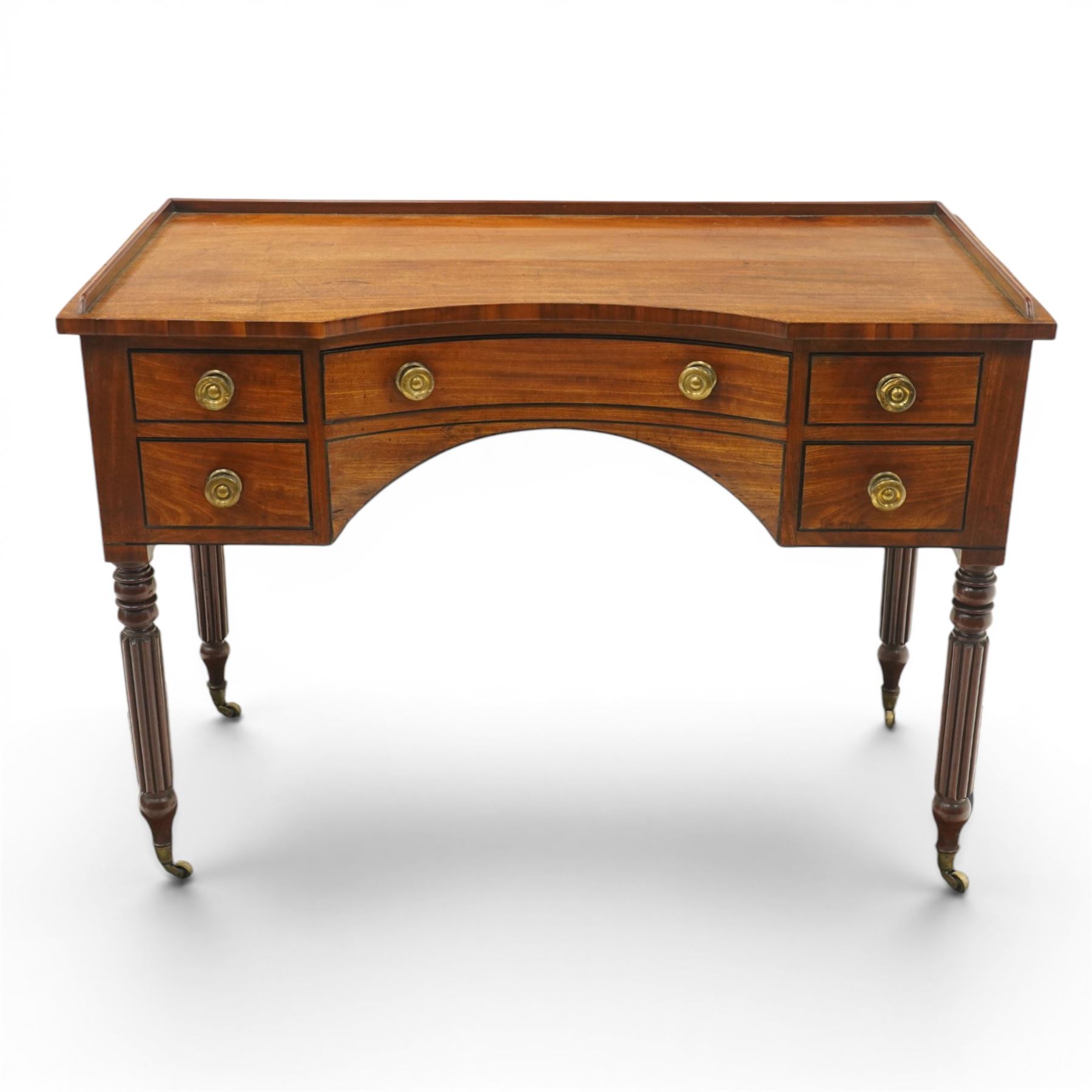 In the manner of Gillows - Regency period mahogany kneehole dressing table or writing desk, the shaped rectangular top with raised three-quarter gallery, fitted with central concave frieze drawer flaked by two graduating drawers to each side, each with ebony stringing, raised on turned and reeded supports terminating to brass cups and castors