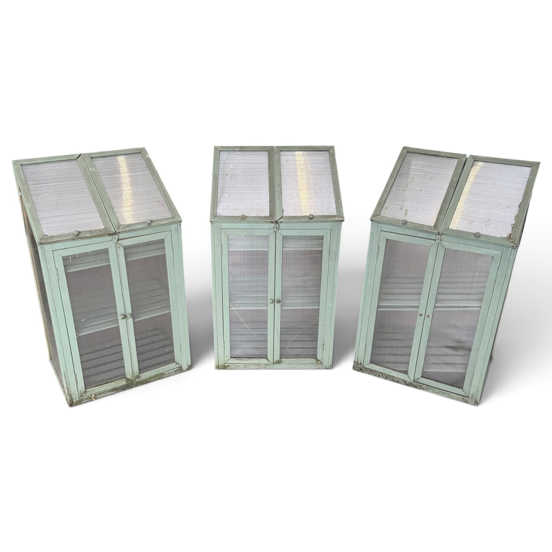 Three green-painted timber cold-frame greenhouses or growhouses, each with twin hinged sloping polycarbonate lids above a pair of panelled doors enclosing slatted shelves