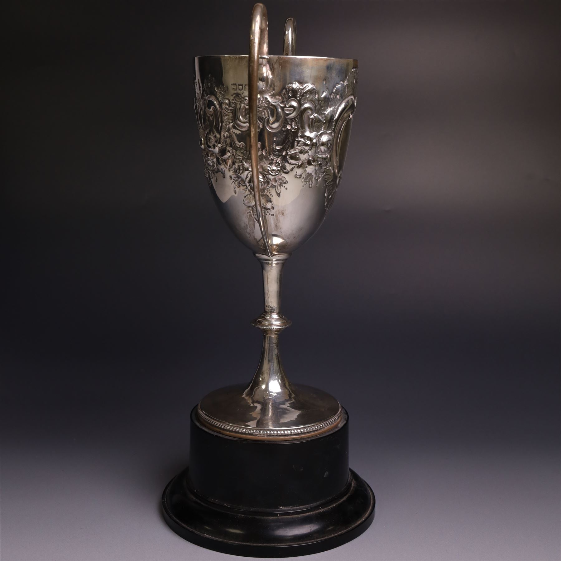 Edwardian silver twin-handled trophy, with embossed floral and scrolling decoration, upon a knopped stem and circular foot, hallmarked Goldsmiths & Silversmiths Co Ltd, London 1903, upon a black plinth, including plinth and handles H32cm