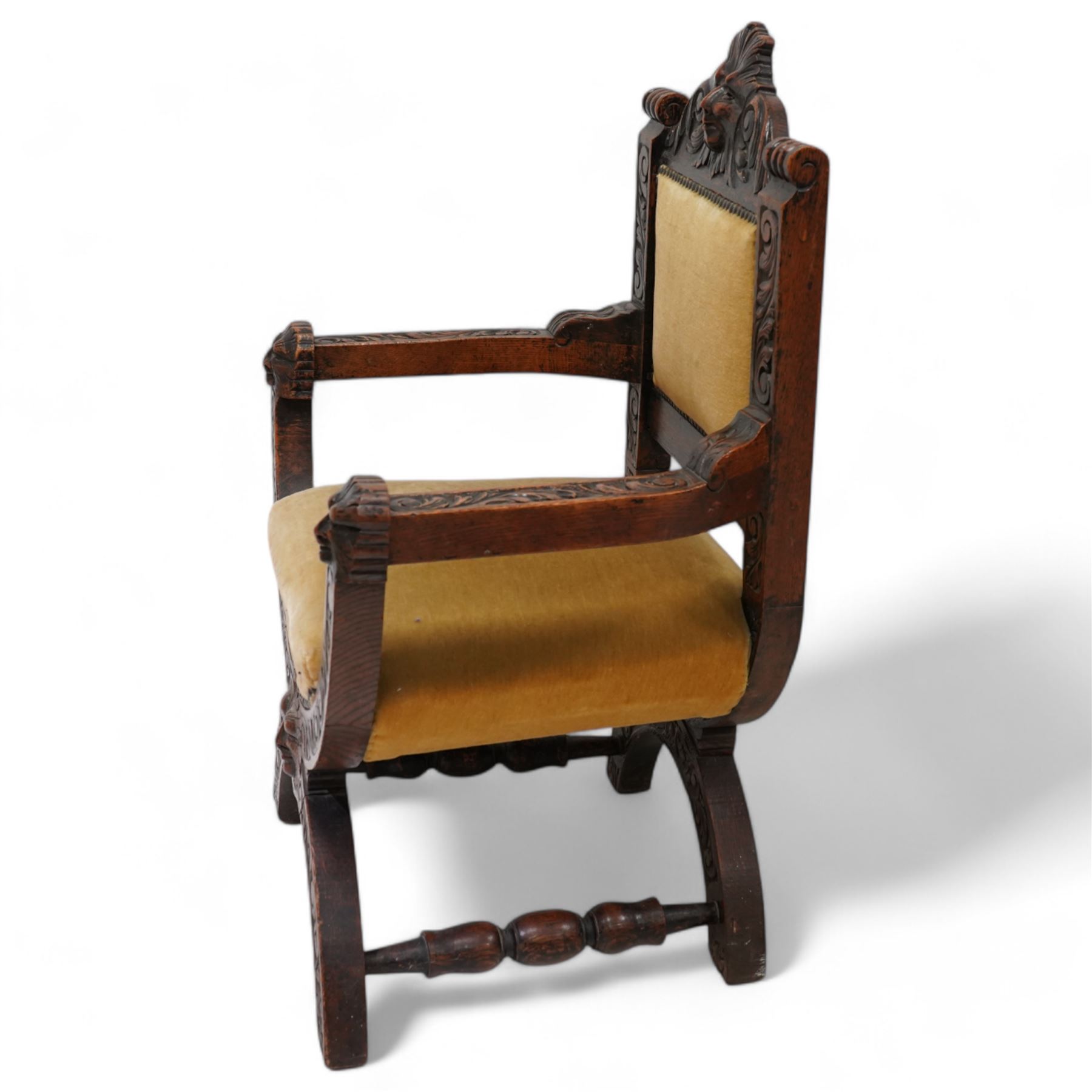Victorian heavily carved oak X-framed throne chair, the cresting rail carved with mask and curling foliage, upholstered in mustard fabric, the arms and frame carved with scrolling foliage, united by turned stretchers 