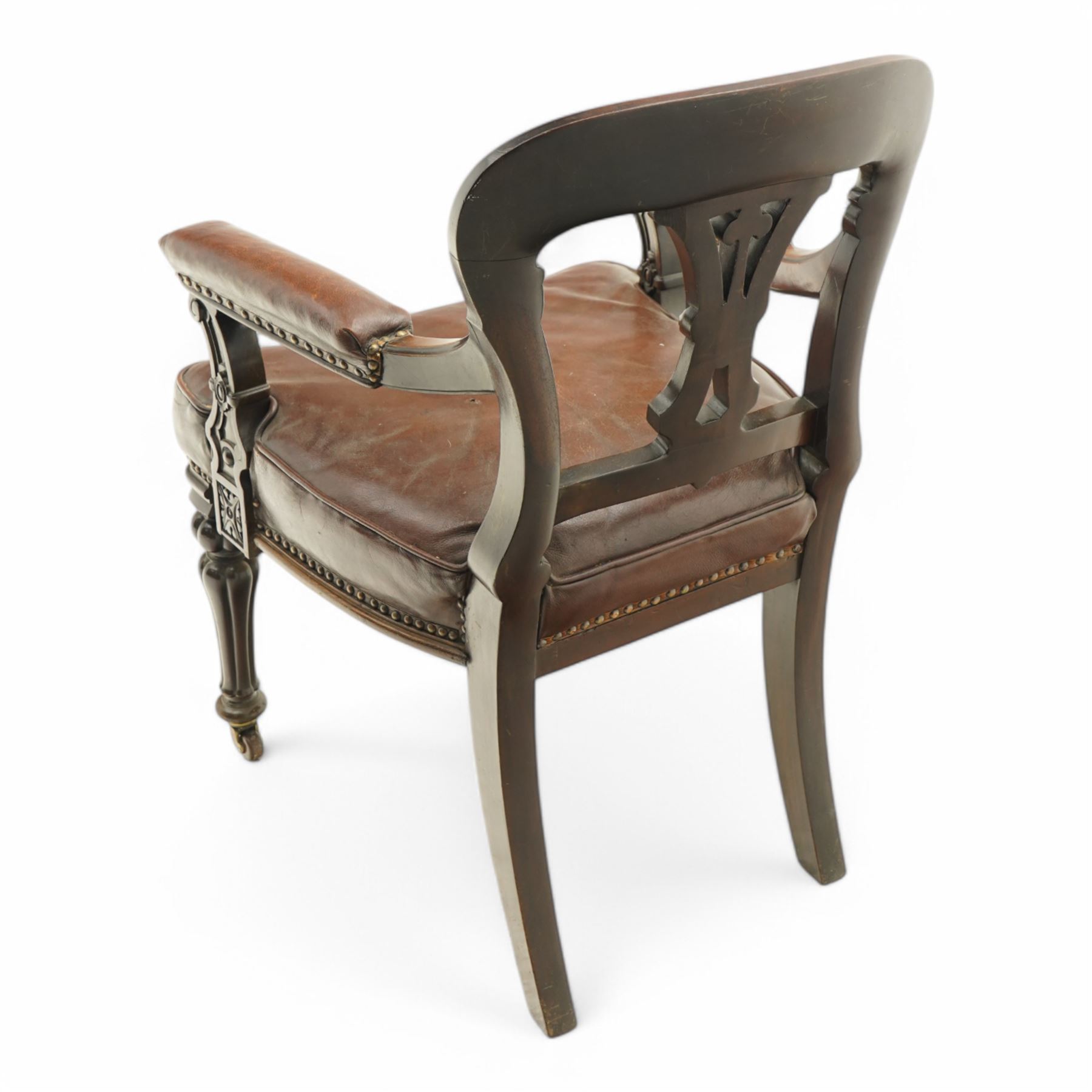Victorian mahogany library elbow chair, arched cresting rail over shaped and pierced splat carved with cartouche, upholstered arms and seat in brown leather with studded bands, turned and lobe carved front supports on brass and ceramic castors 