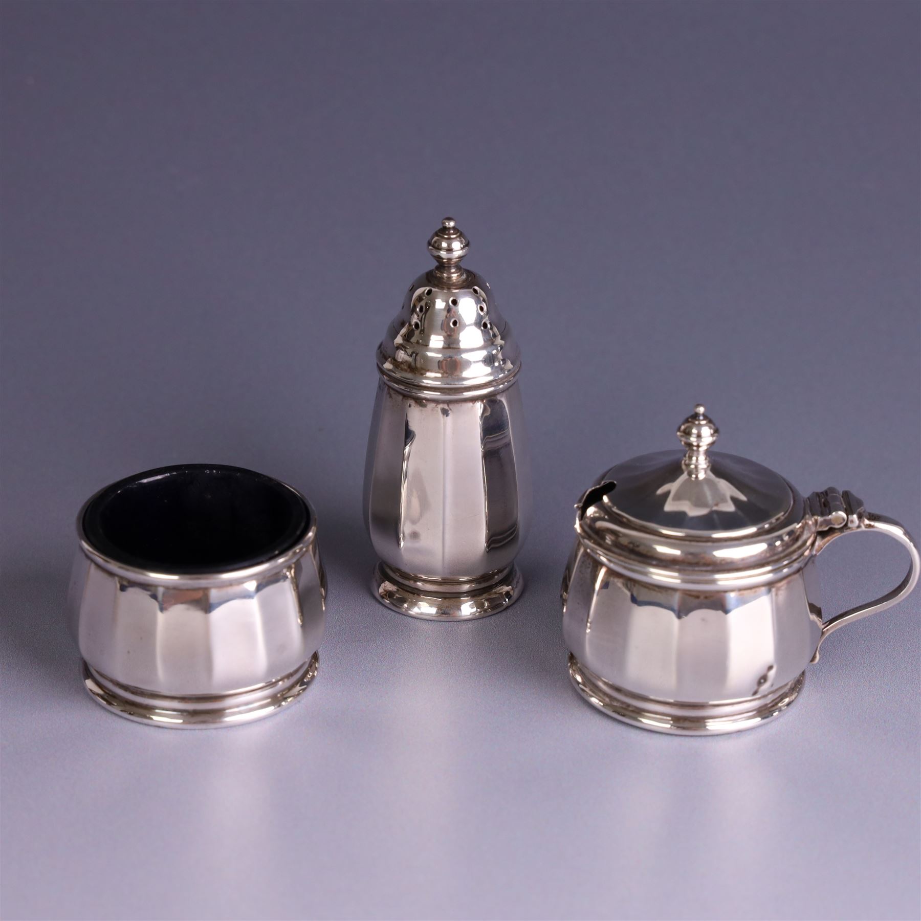 Mid 20th century three piece silver cruet set, comprising pepper shaker, open salt and mustard pot with cover, each of circular faceted form, hallmarked Walker & Hall, Birmingham 1958, the mustard pot and salt with blue glass liners