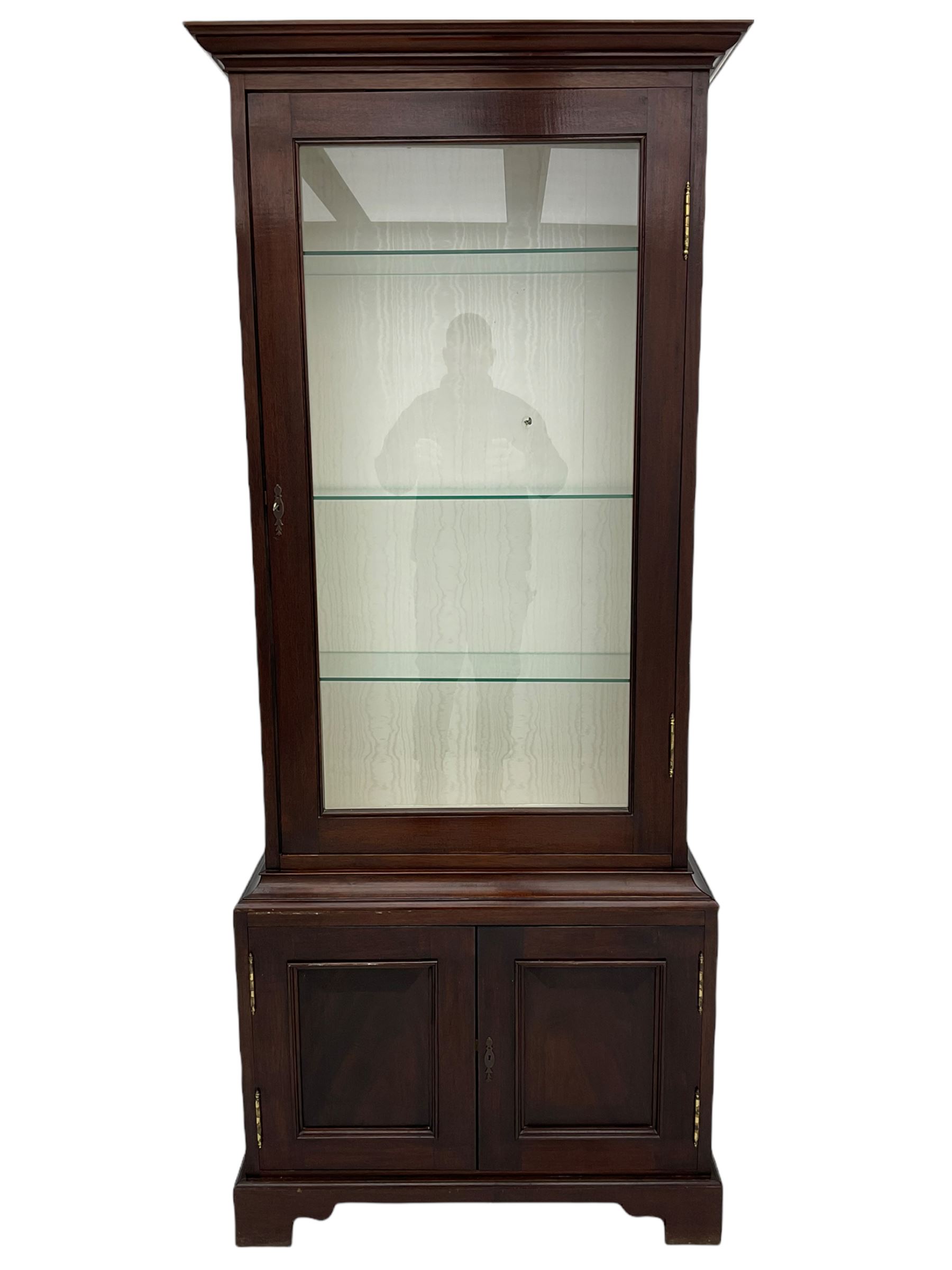Mahogany display cabinet with hidden compartment, projecting cornice over single glazed door enclosing glass shelves, double panelled cupboard below, the interior with button releasing sliding back compartment, on bracket feet