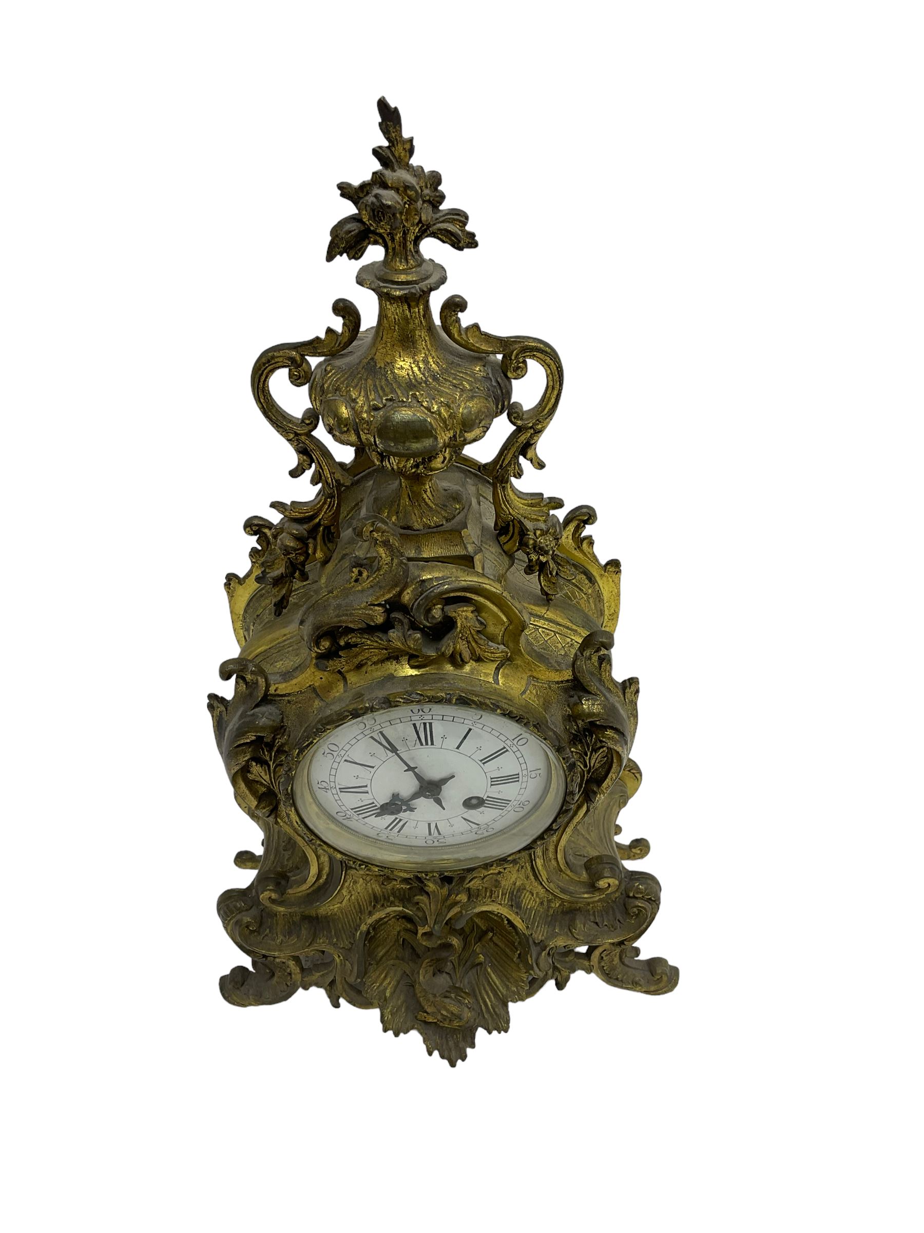 French - late 19th century 8-day gilt brass mantle clock surmounted by an urn, with a waisted case and baroque decoration raised on four splayed feet, with a glazed bezel, enamel dial with Roman numerals, five minute Arabic's and gothic steel hands, count wheel striking movement, striking the hours and half-hours on a bell. With pendulum.