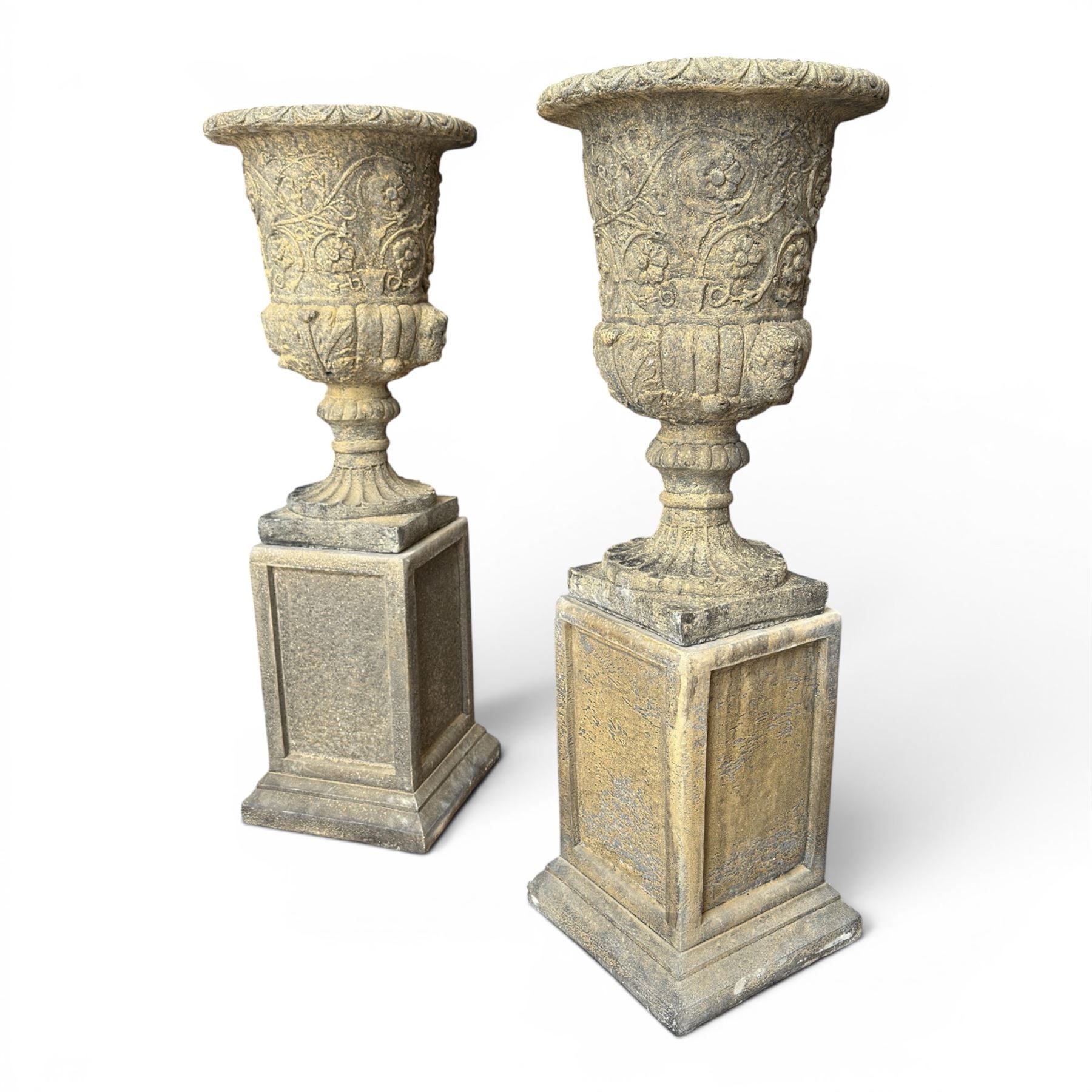 Pair of George III design antiqued composite stone garden urns of campana form, the egg-and-dart everted rim over a waisted body relief moulded with scrolling fruiting foliage and flowerheads, the gadrooned underbelly with twin mask ornaments, the knopped stems on fluted socles, raised on faux panelled square pedestal bases