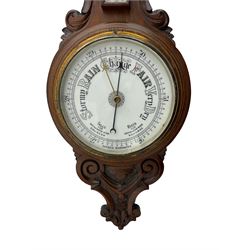 An English 1930’s solid oak carved hall barometer in a scroll shaped carved case with relief carving, aneroid movement, 8” white register measuring barometric air pressure from twenty-six to thirty-one point nine inches, weather predictions in black upper and lower case gothic script with a blue steel indicating hand and brass recording hand, brass dial bezel with flat bevelled glass, mercury thermometer enclosed in a glazed rectangular box displaying temperature in degrees Fahrenheit and Celsius on a white register. H87cm



