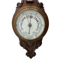 An English 1930’s solid oak carved hall barometer in a scroll shaped carved case with relief carving, aneroid movement, 8” white register measuring barometric air pressure from twenty-six to thirty-one point nine inches, weather predictions in black upper and lower case gothic script with a blue steel indicating hand and brass recording hand, brass dial bezel with flat bevelled glass, mercury thermometer enclosed in a glazed rectangular box displaying temperature in degrees Fahrenheit and Celsius on a white register. H87cm


