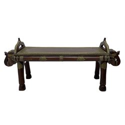 Indian hardwood and brass bound bench or coffee table, the rectangular top with embossed brass banding in a repeating flower head pattern, flanked by pierced handles, raised on twin end supports in the form of elephant masks decorated with brass-work, over elephant leg supports