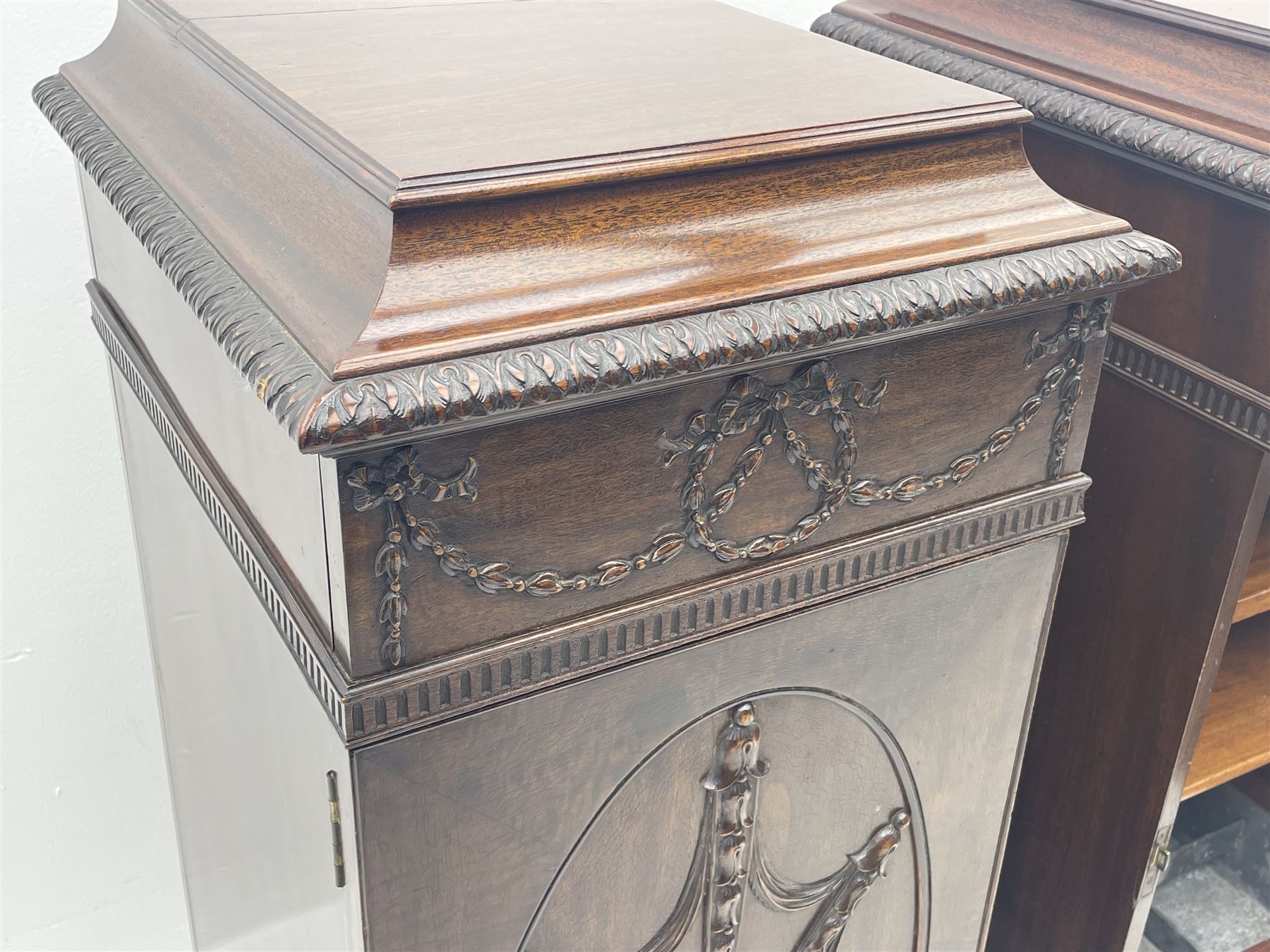 Pair early 20th century Hepplewhite style mahogany pedestal cabinets, cavetto moulded top with foliate carved edge over secret frieze drawer with applied trailing floral and ribbon decoration, each with button release underneath, oval panelled door with trailing harebell and central flower head motif decoration enclosing shelves, fitted cutlery drawer and lined cellarette drawer, on turned acanthus carved feet