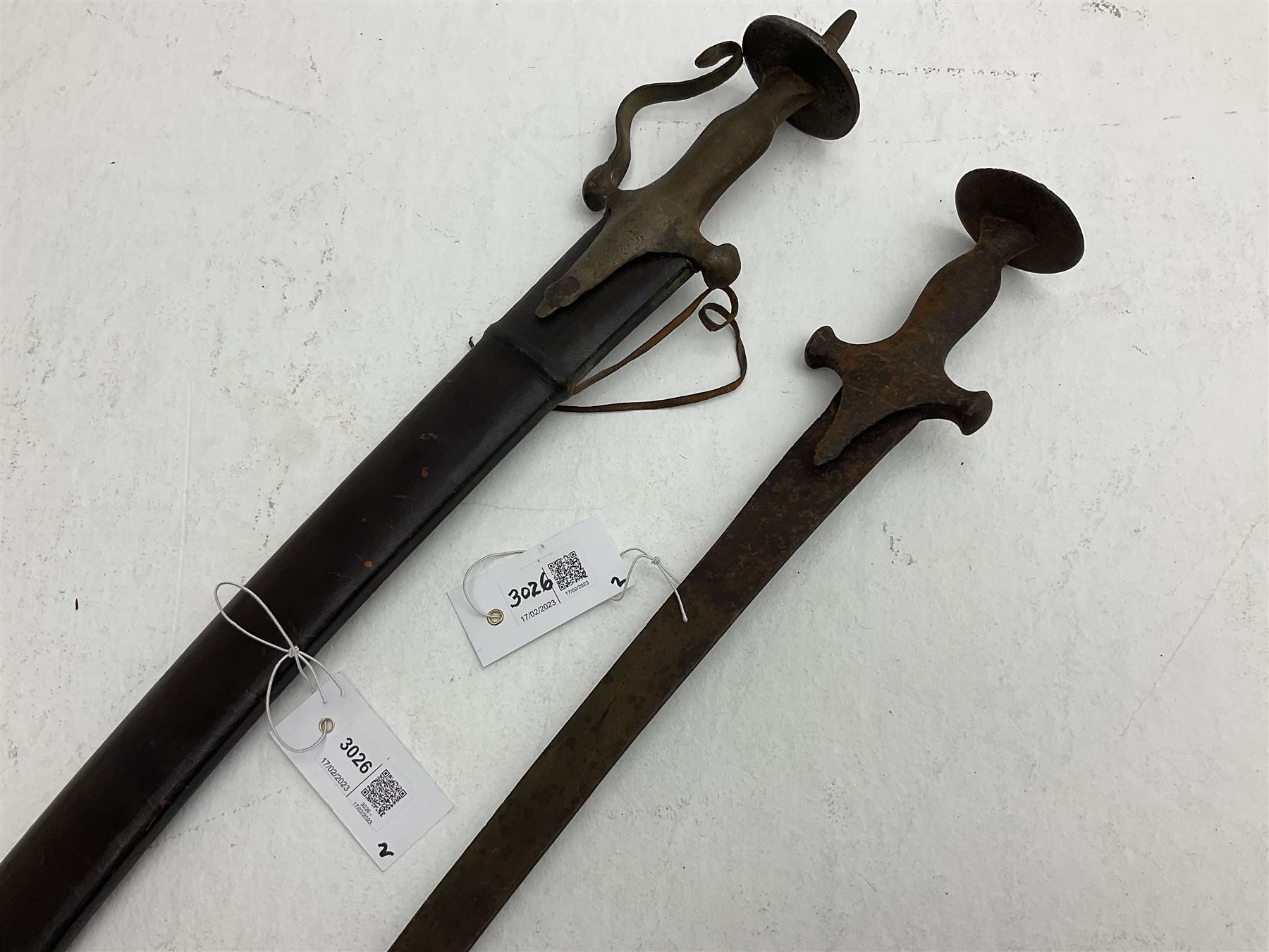 Late 19th/early 20th century Indian Tulwar sword with 81cm plain curving steel blade and iron hilt with extended langets, knucklebow and spiked pommel; in leather covered scabbard L100cm overall; and another Tulwar lacking scabbard in almost relic condition (2)