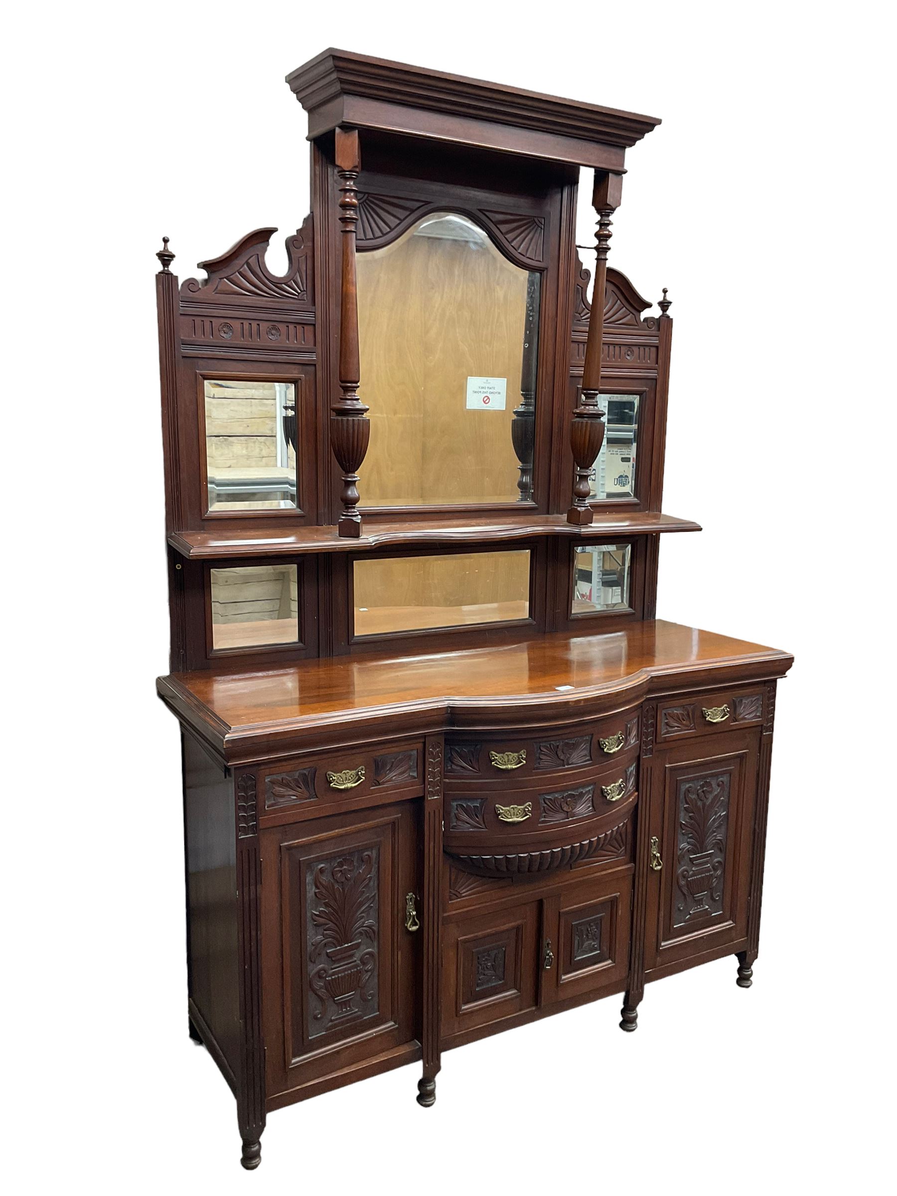 Edwardian walnut sideboard, projecting cornice supported by turned supports, the back fitted with three bevelled mirrors beneath fluted and flowerhead carved friezes, the lower section with four drawers and four panelled cupboard doors carved with foliate and urn designs, fluted uprights on turned feet
