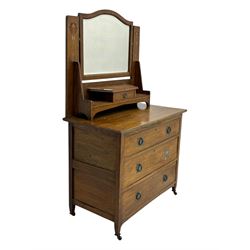 Early 20th century Arts and Crafts oak dressing chest, the arched mirror flanked by floral marquetry inlaid uprights above a shelf and drawer, over three long drawers with square hammered metal plates and circular pulls, on slender supports with castors