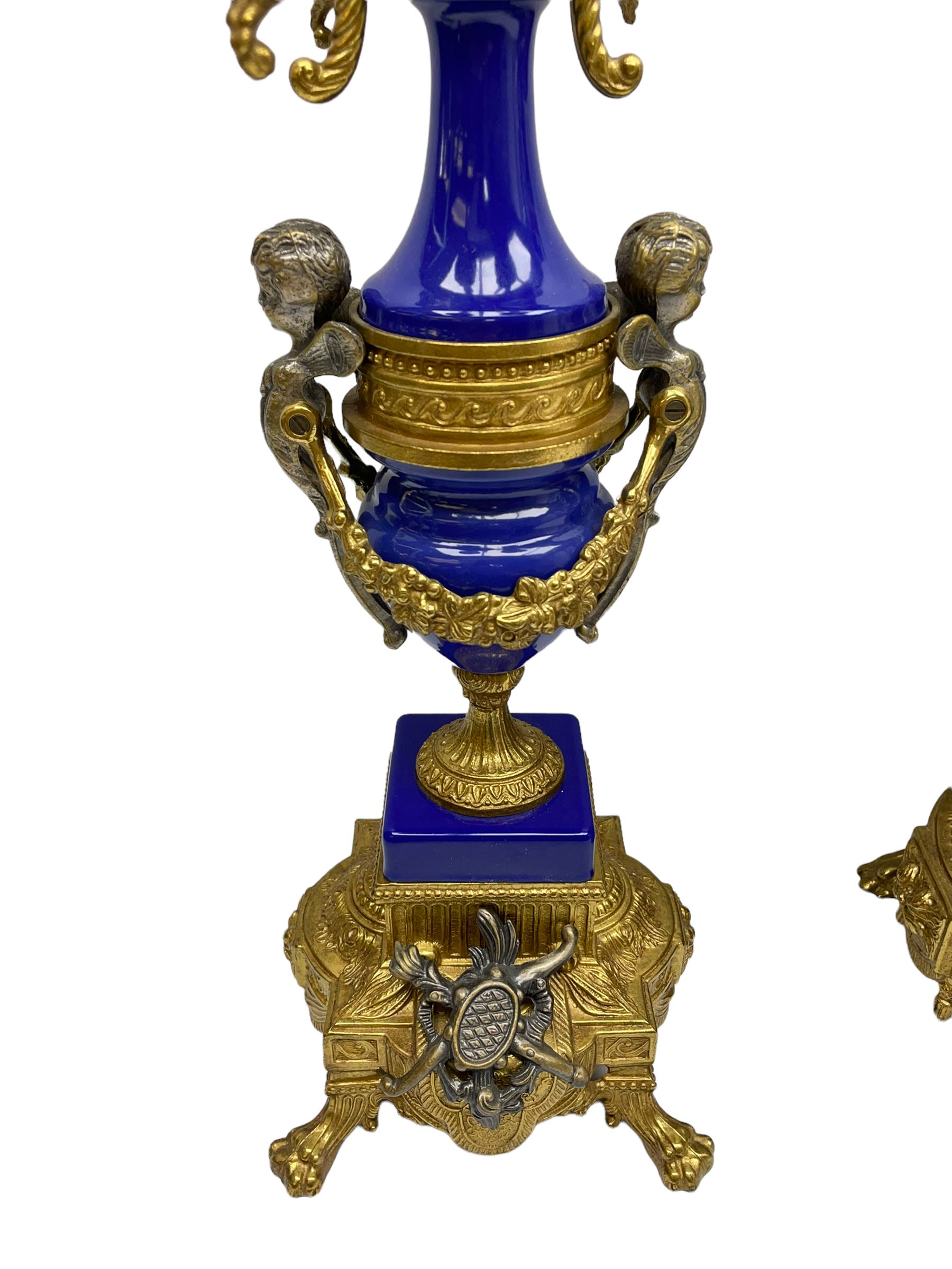 A 20th century continental gilt metal Lyre mantle clock with a pair of matching six light candelabra, spring driven movement housed in a gilt drum case on a brass and cobalt blue base with paw feet, two bronzed metal figures of mythological cherubs with animal legs and cloven hooves supporting a festooned garland swag, eight-day twin barrel striking movement with a floating lever balance escapement, striking the hours and half-hours on two bells, white enamel dial with roman numerals, minute track and pierced steel hands, dial inscribed “Imperial”. 