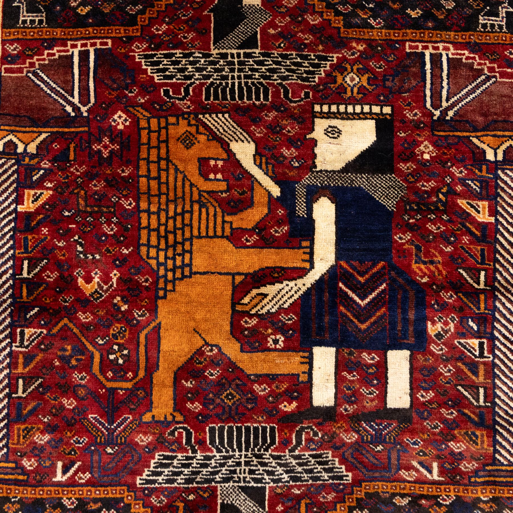 South West Persian Qahqai pictorial hand-knotted rug, madder red field with an all-over pattern of repeating rosettes enclosed with angular leaves and corners with stylized animals figures, central stylized lion in rearing stance and raised forelegs and a human figure shown in profile within a principal border of alternating octagonal and diamond medallions, each enclosing stylized rosettes, flanked by guard stripes incorporating latch hook motifs, with cotton fringe ends  