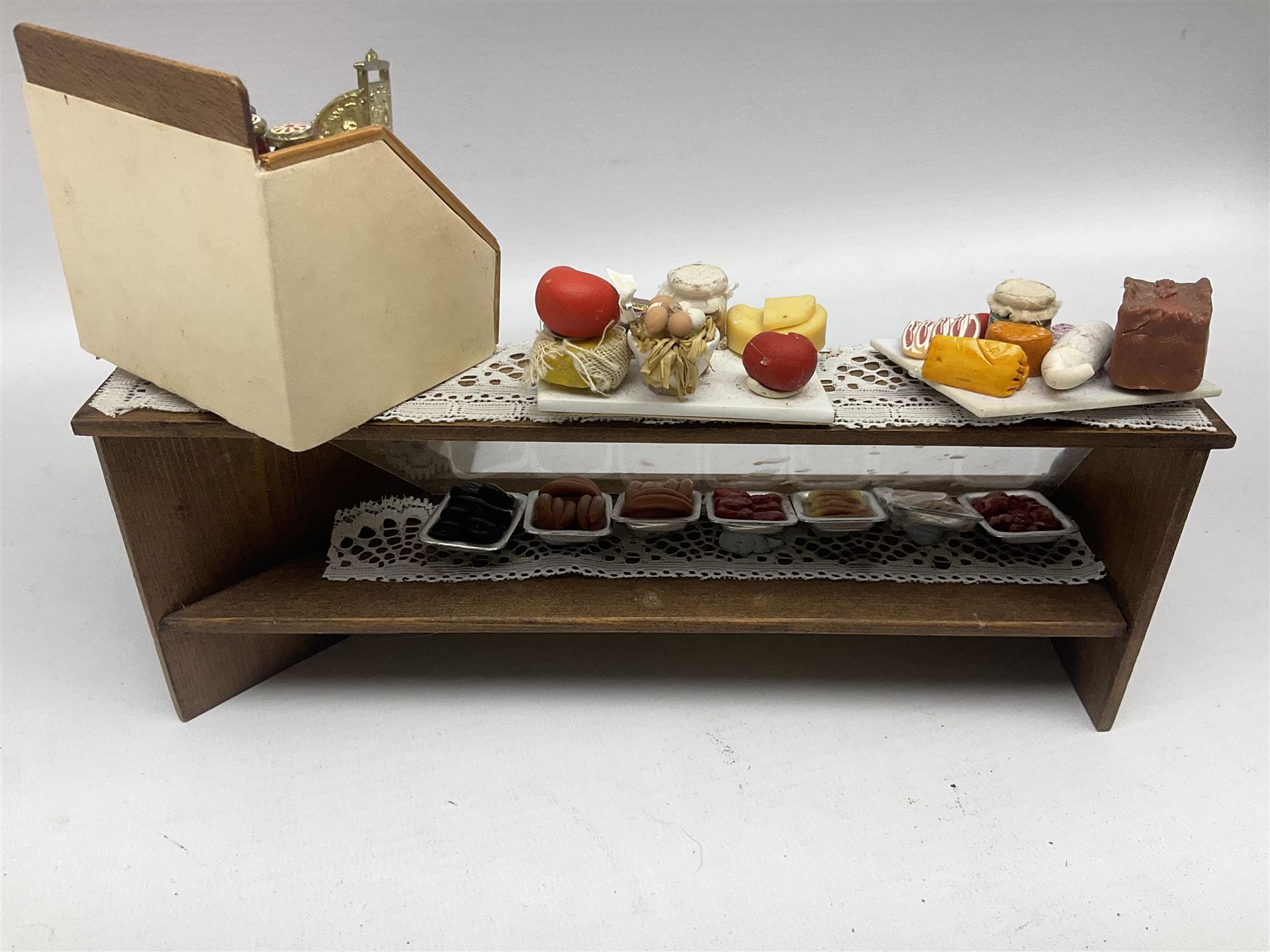 Collection of miniature dolls house grocery shop furniture and produce, comprising glazed corner counter with brass style cash register, shelved preserves unit, pies, cheese board and trays of meats, pair of white cheese counters, biscuit display with six tins, glazed preserves cabinet, pair of shop chairs and four further cheese boards complete with grapes and label flags