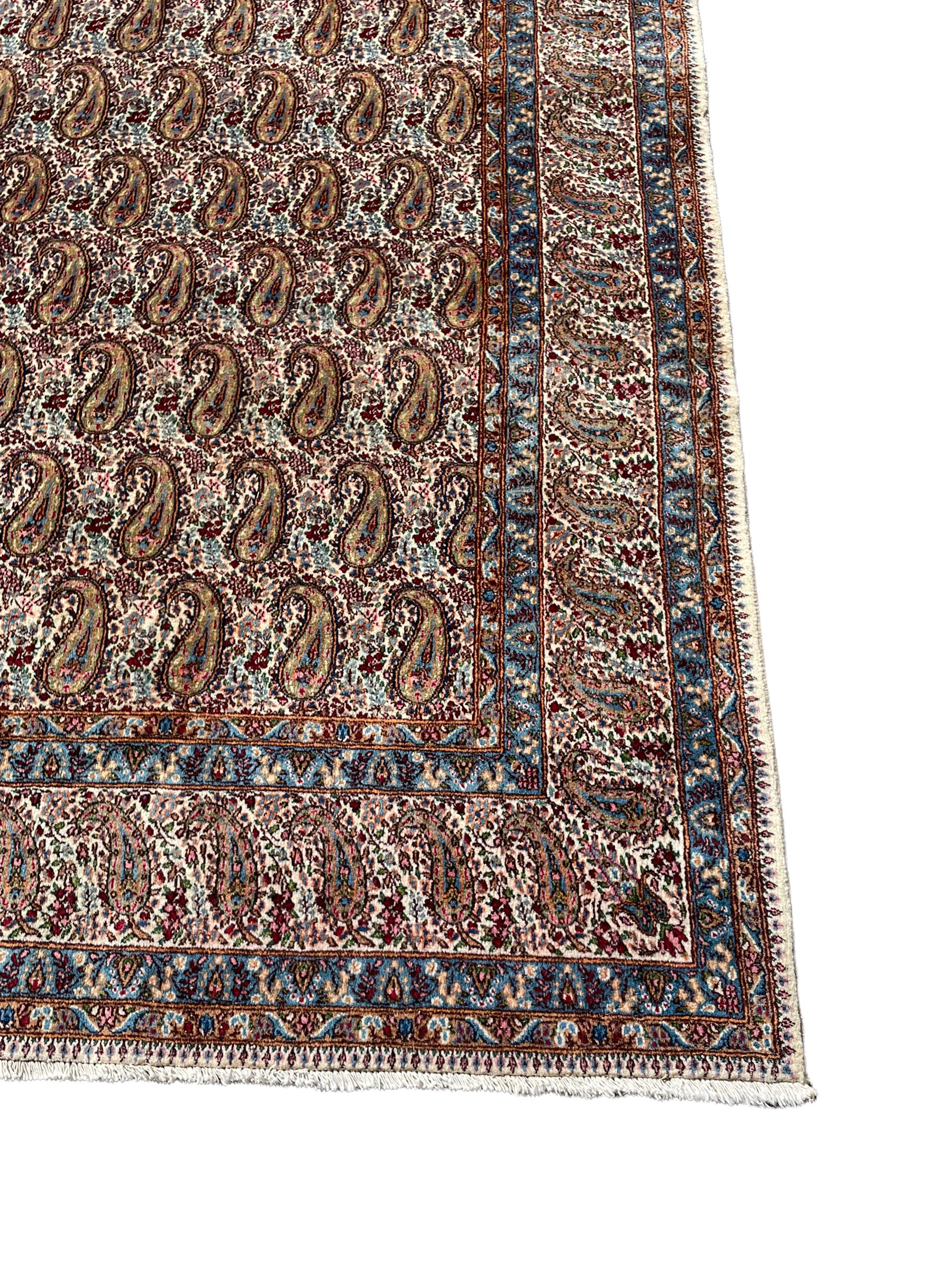 Persian Kirman carpet, the ivory ground field and border decorated with repeating Boteh motifs, interspaced with all-over floral design, blue ground border guards, the top border with signature panel