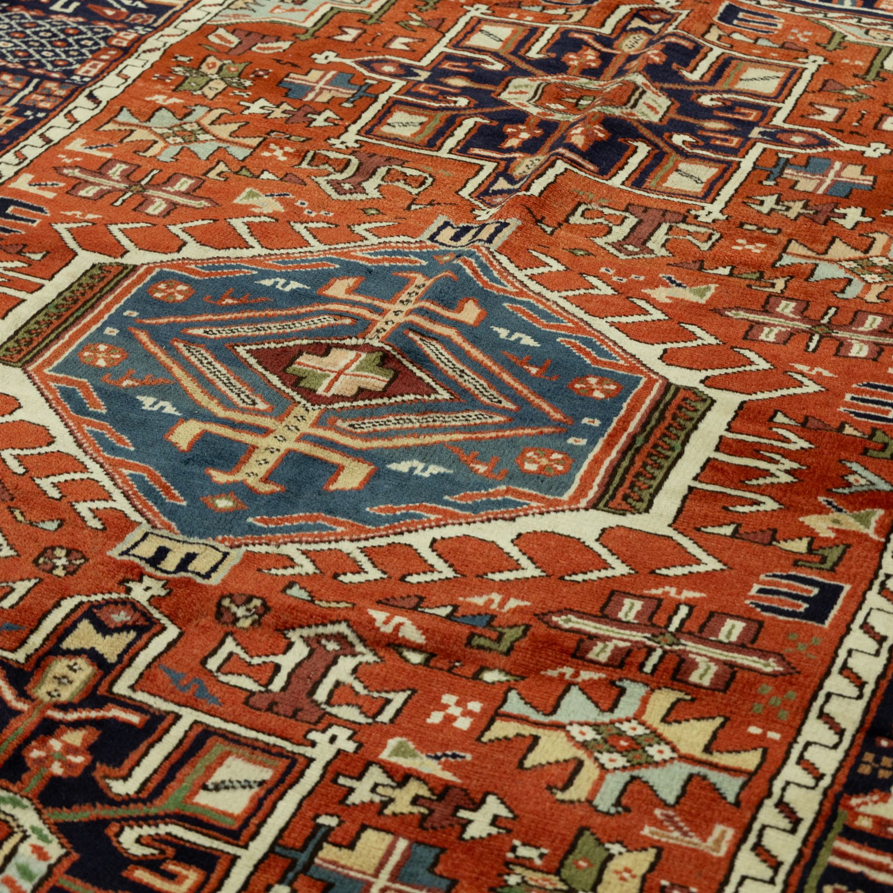North West Persian Karajeh hand-knotted rug, terracotta ground, with a stepped geometric pole medallion design in blue, ivory and dark navy tones, surrounded by angular tribal motifs incorporating stylised flowerheads, hooked devices and geometric vine work, enclosed by a dark navy border with repeating rosettes and angular leaf motifs, flanked by narrow patterned guard stripes, with cotton fringes