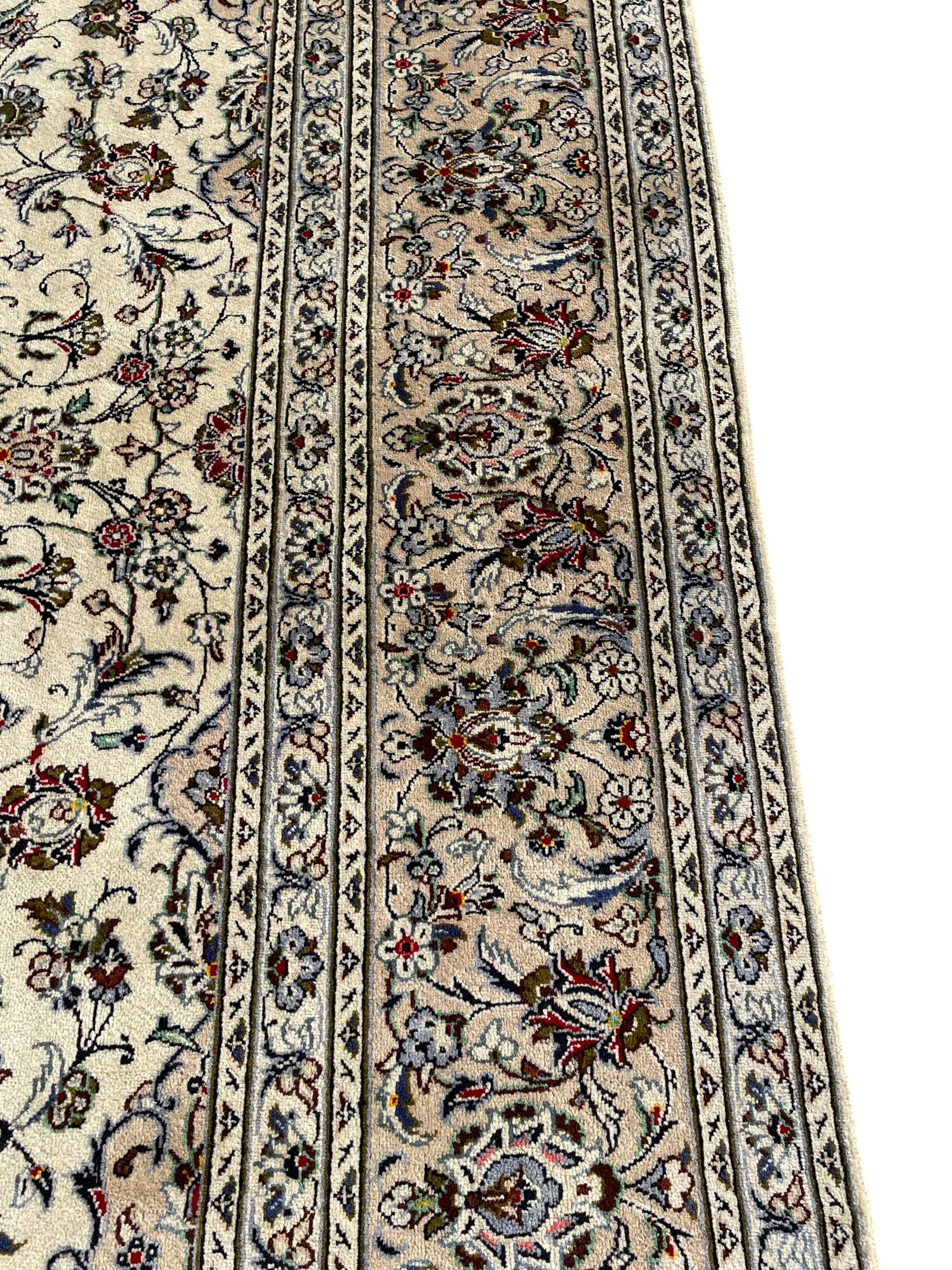 Persian Kashan ivory ground carpet, central rosette medallion with projecting palmettes, densely decorated field depicting trailing leafy branches and stylised plant motifs, the border with similar scrolling pattern, within floral pattern guard stripes 