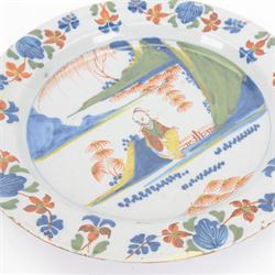 Mid 18th century Lambeth Delft plate, probably from the Abigail Griffith factory, circa 1750, painted in polychrome enamels with a figure seated in a landscape, within a garlanded border of flowers to rim, D34cm, together with a Liverpool Delft plate, circa 1760, painted in blue with a boathouse surrounded by trees by a river, within diaper and cell borders, D37cm