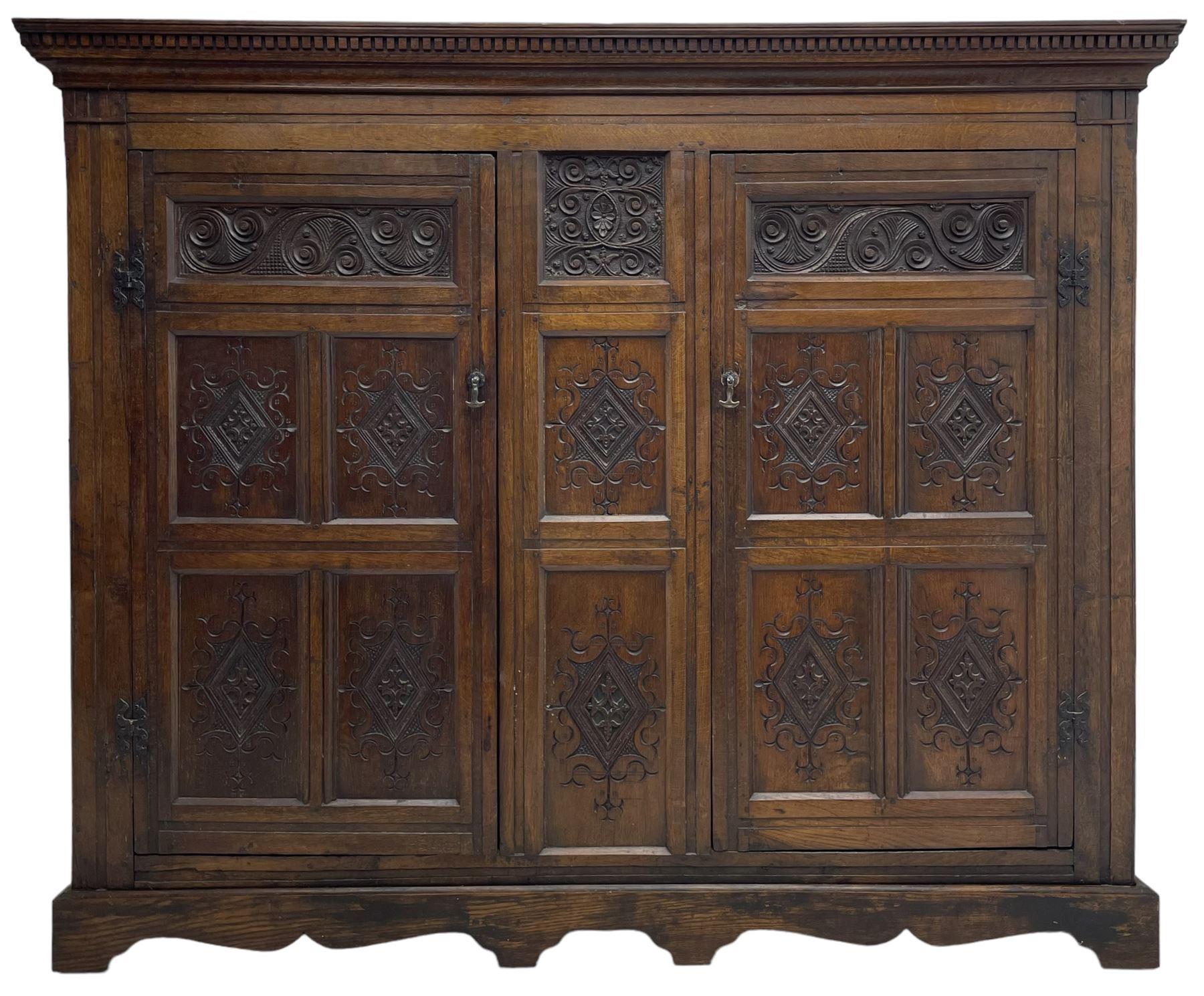 Large 18th century and later oak livery cupboard, projecting moulded and dentil cornice, sunken fillet moulded frame, triple vertical panelled central upright, enclosed by two doors each with five panels, the top row of panels carved with stylised scrolls, the lower panels carved with foliate lozenges, on shaped bracketed skirt base 