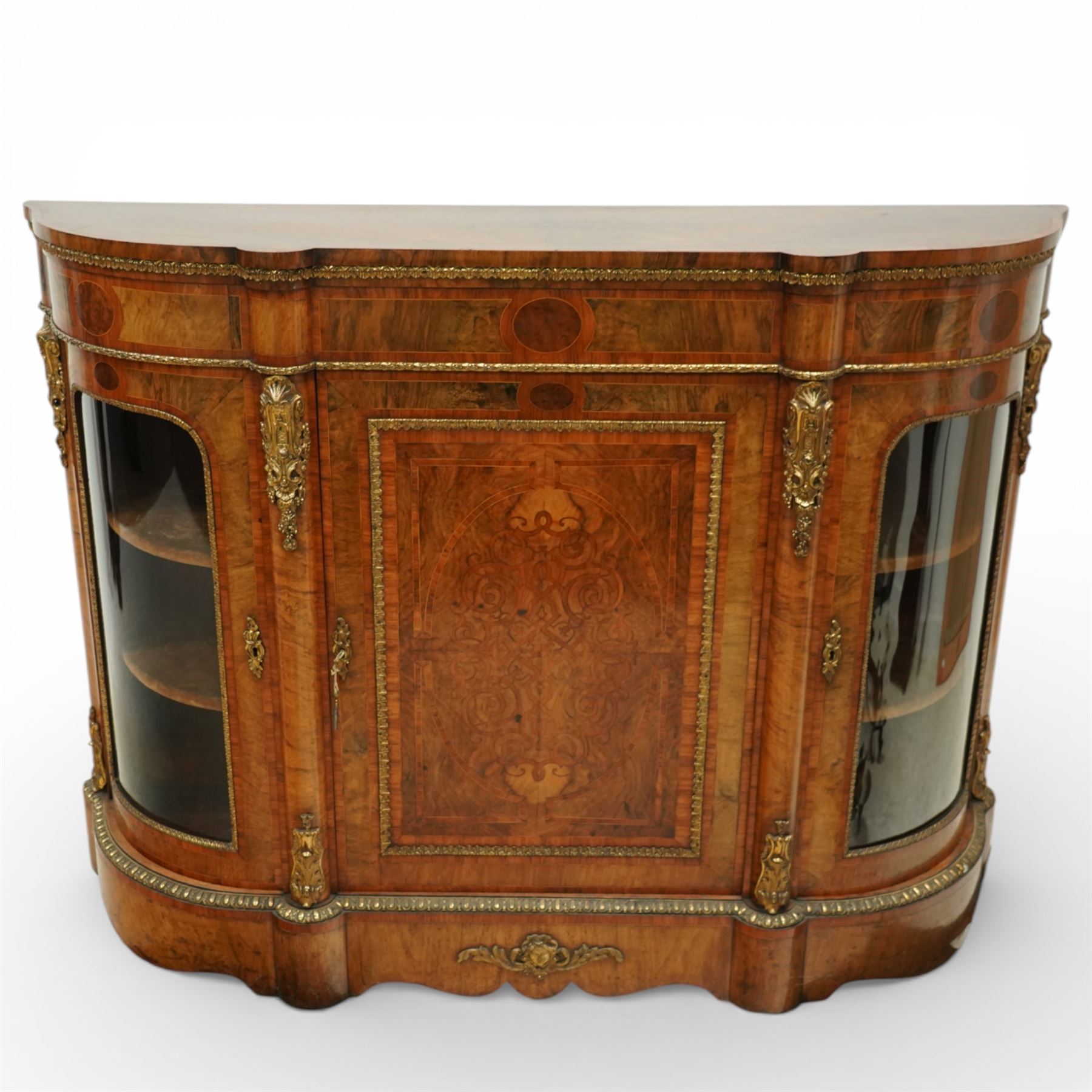Late Victorian figured walnut credenza, the shaped demi-lune top with cross-banding and stringing over an inlaid frieze with foliate gilt-metal mounts, fitted with a central geometric-inlaid panelled door, flanked by convex-glazed side-cabinets with a gilt metal slip and outer-banding, the uprights surmounted by gilt-metal cartouche mounts with cascading foliage details, raised on a shaped skirted base with central female-mask gilt mount