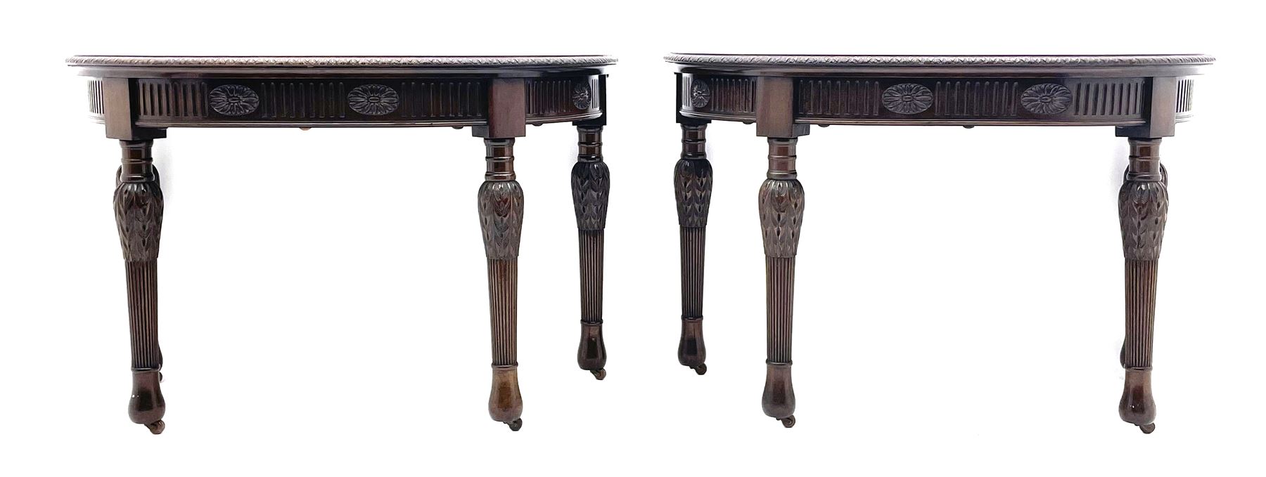 Pair early 20th century Hepplewhite style mahogany D-end console tables, the curved top with foliate carved edge, fluted frieze rails carved with flower heads, turned and reeded acanthus supports with recessed castors