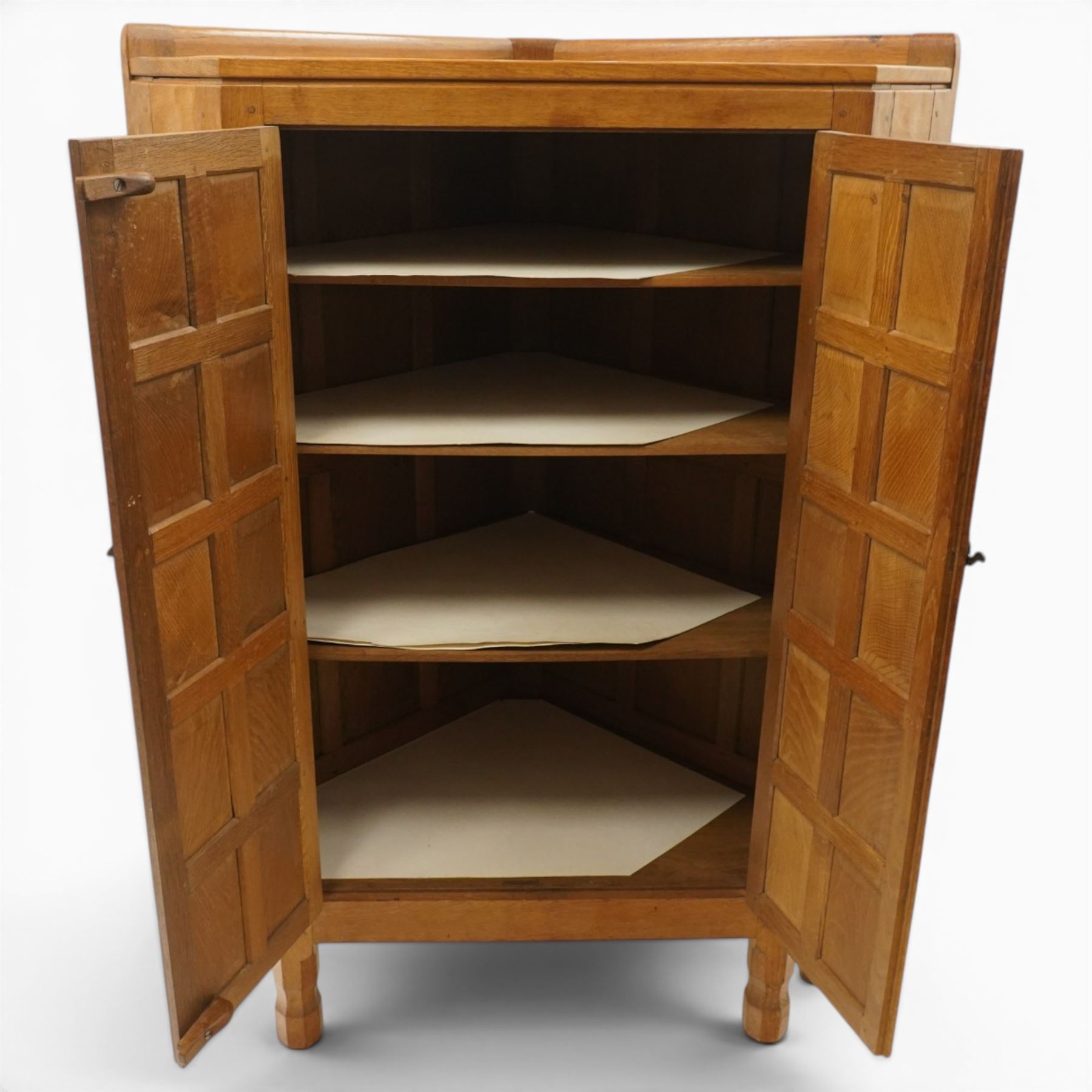 Mouseman - 1940's adzed and panelled oak corner cupboard, the raised quarter-gallery back over twin adzed panelled doors with wrought iron fittings and latch, enclosing three shaped shelves, raised on turned octagonal supports, with carved mouse signature in relief, by the workshop of Robert Thompson of Kilburn Provenance: purchased directly from the Kilburn workshop in the late 1940s