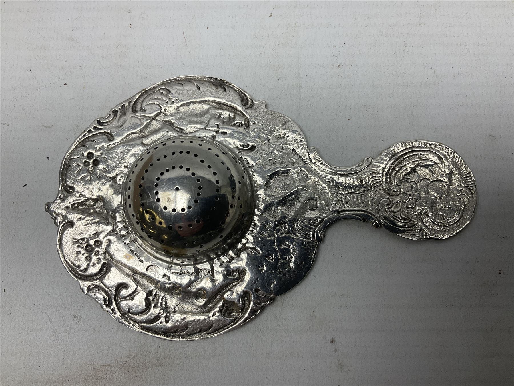 Continental white metal strainer, of shaped form embossed with classical figures, putto, musical trophies and fruiting vines, indistinctly marked, L12.5cm