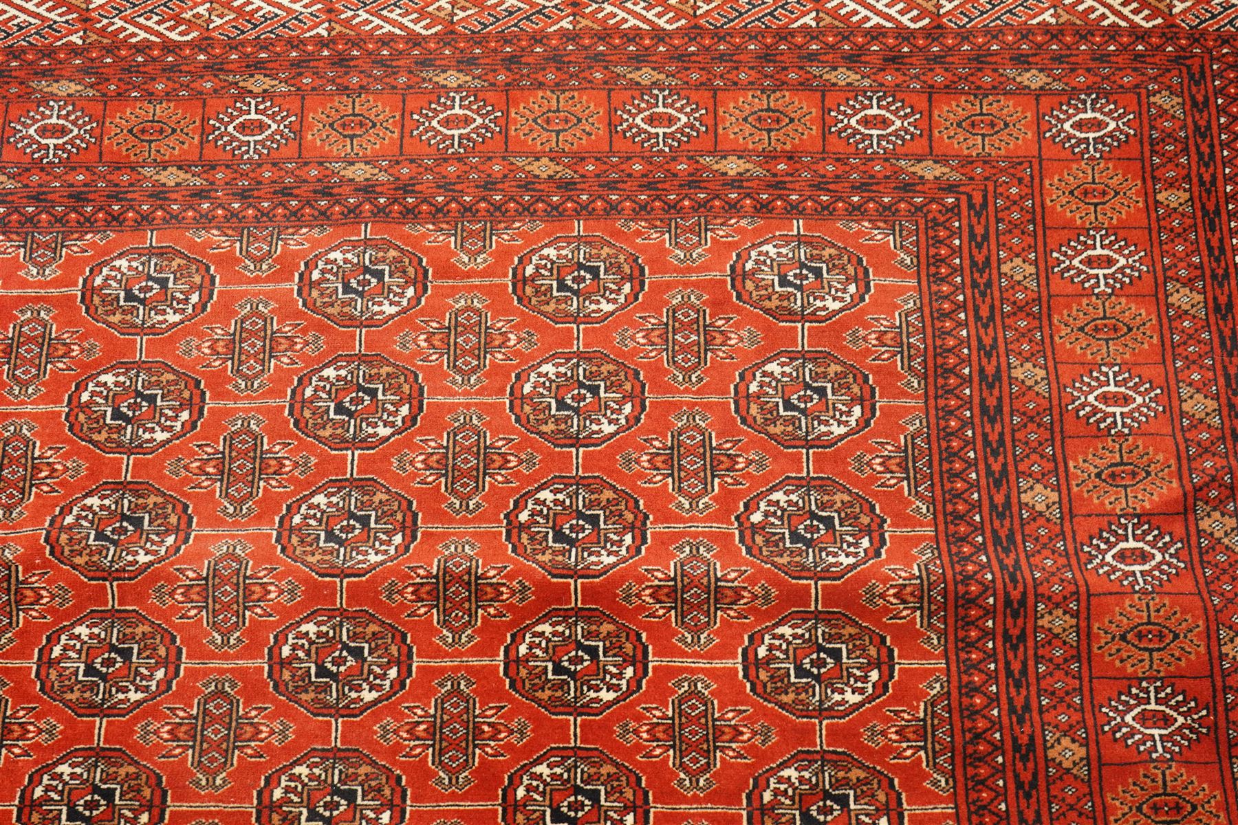 Turkmen hand-knotted carpet, madder red field, with an all-over design of row of Tekke guls, each enclosing stylized cruciform and geometric devices in tones of ivory, dark brown, and indigo, principal border of stylized geometric motifs, flanked by guard border with angular latch hook geometric designs, with cotton fringe ends 