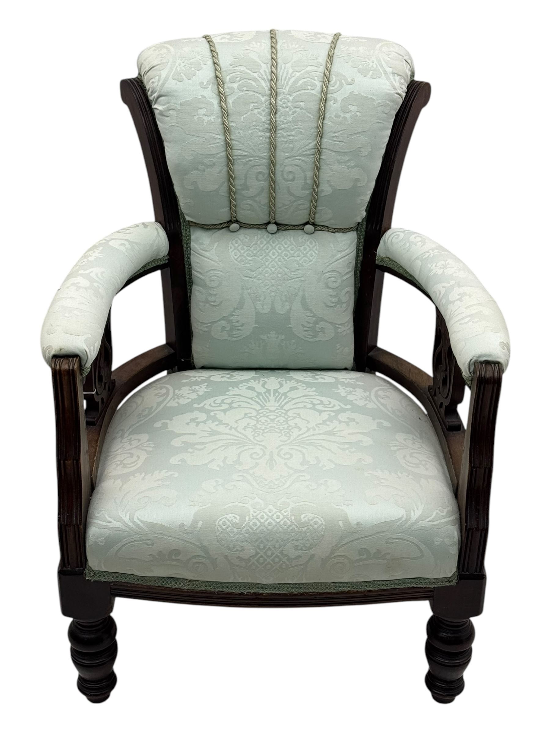 Late Victorian carved mahogany open armchair, the shaped back with fluted upholstered panel and padded arms above pierced scroll carved side supports, over a broad upholstered seat, on turned front supports