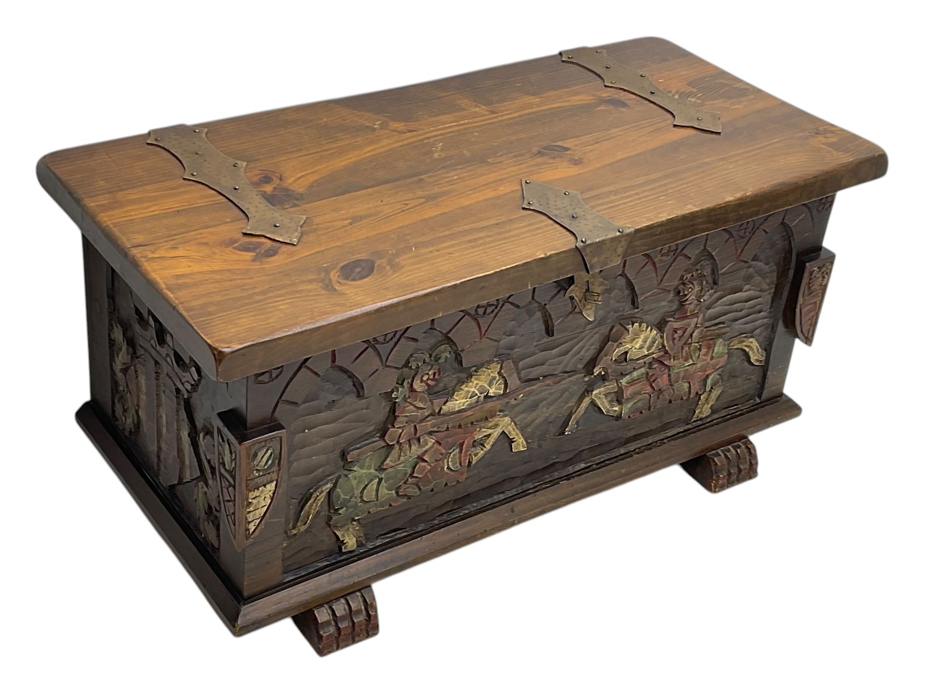 Mid-20th century carved hardwood chest, the rectangular hinged top with decorative strap hinges, the front and sides deeply carved with medieval-style jousting knights, heraldic shields, and architectural elements, raised on shaped block feet