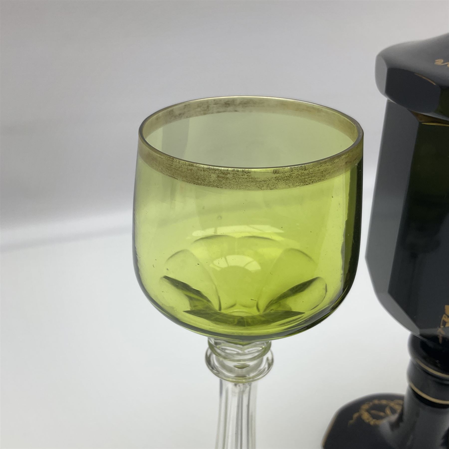 19th Century Bohemian amethyst glass lidded goblet, the bowl of faceted form, gilded with three children in period dress playing games, upon a knopped stem and conforming faceted foot, together with an art nouveau green glass goblet, with gilded rim, upon clear trumpet stem, and a hand blown wine glass, transfer printed with birds and three figures in period dress, tallest H29cm