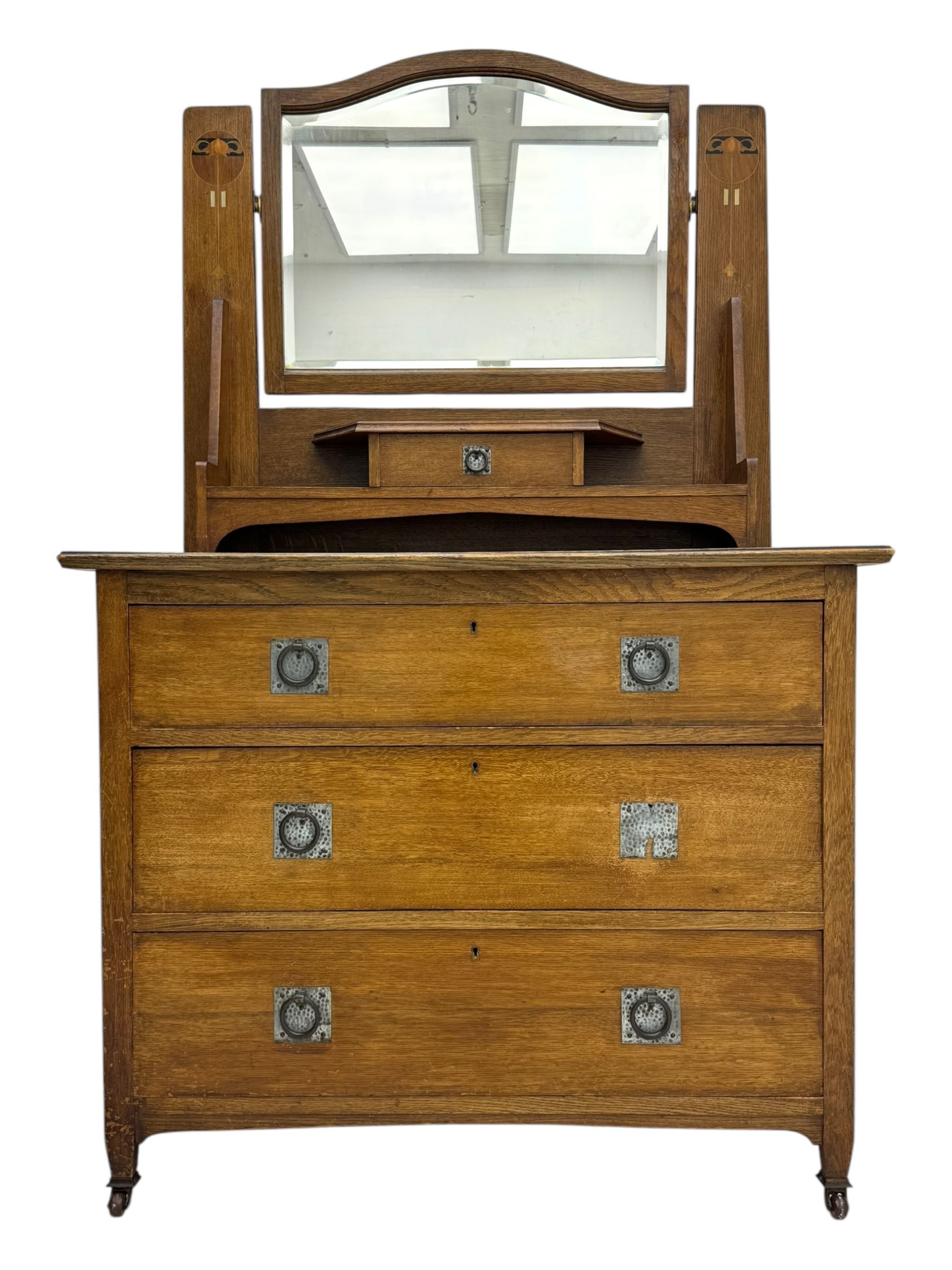 Early 20th century Arts and Crafts oak dressing chest, the arched mirror flanked by floral marquetry inlaid uprights above a shelf and drawer, over three long drawers with square hammered metal plates and circular pulls, on slender supports with castors