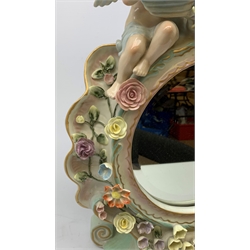  A late 19th century Sitzendorf porcelain mirror, the mirror plate of circular form set within a shaped surround encrusted with flowers and foliage, surmounted by three figures of cherubs and raised upon four scroll feet, with blue cross hatch mark beneath, H57cm.   