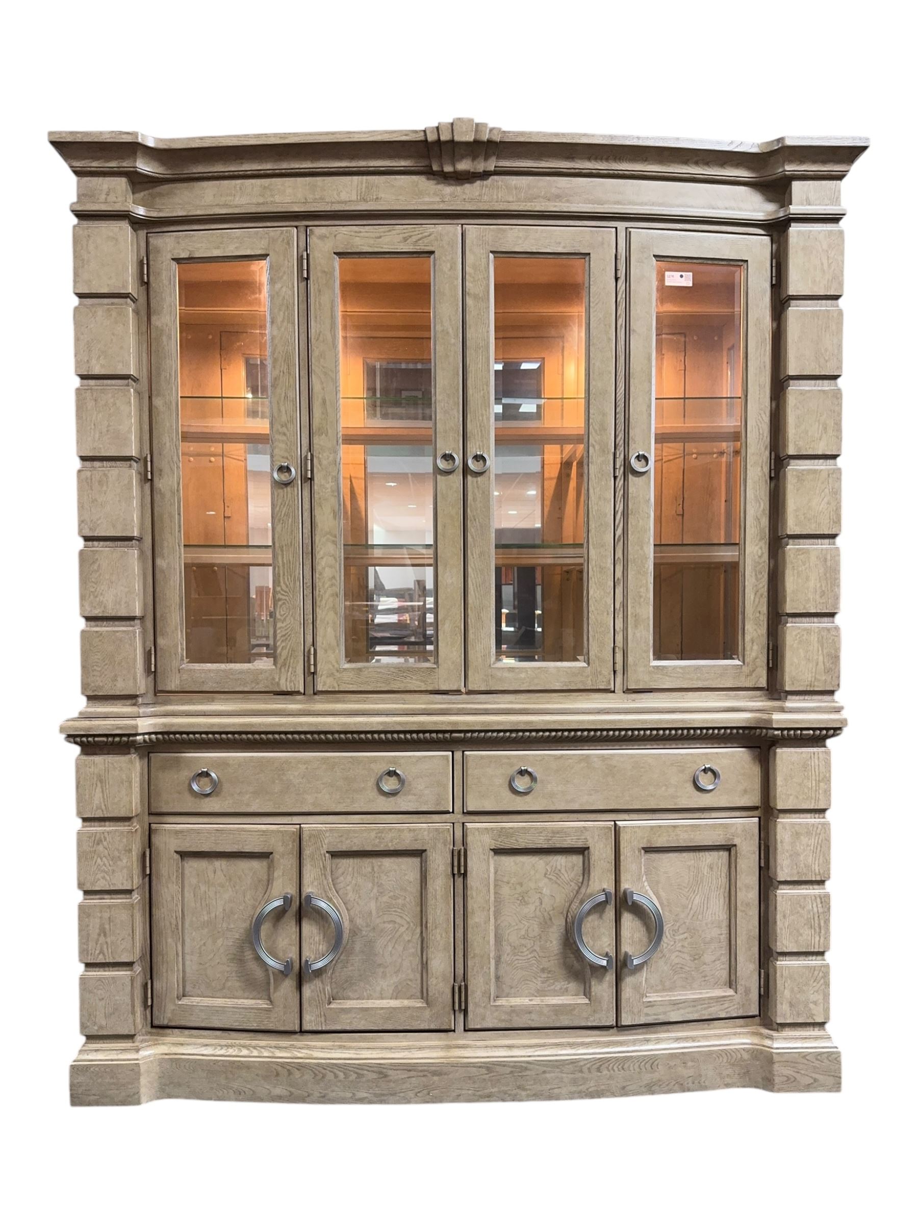 Brunswick - washed oak finish illuminated display cabinet, upper section with four glass-panelled doors enclosing glass shelving, base fitted with two drawers over four paneled cupboard doors, each fitted with distinctive round metal handles, raised on plinth base