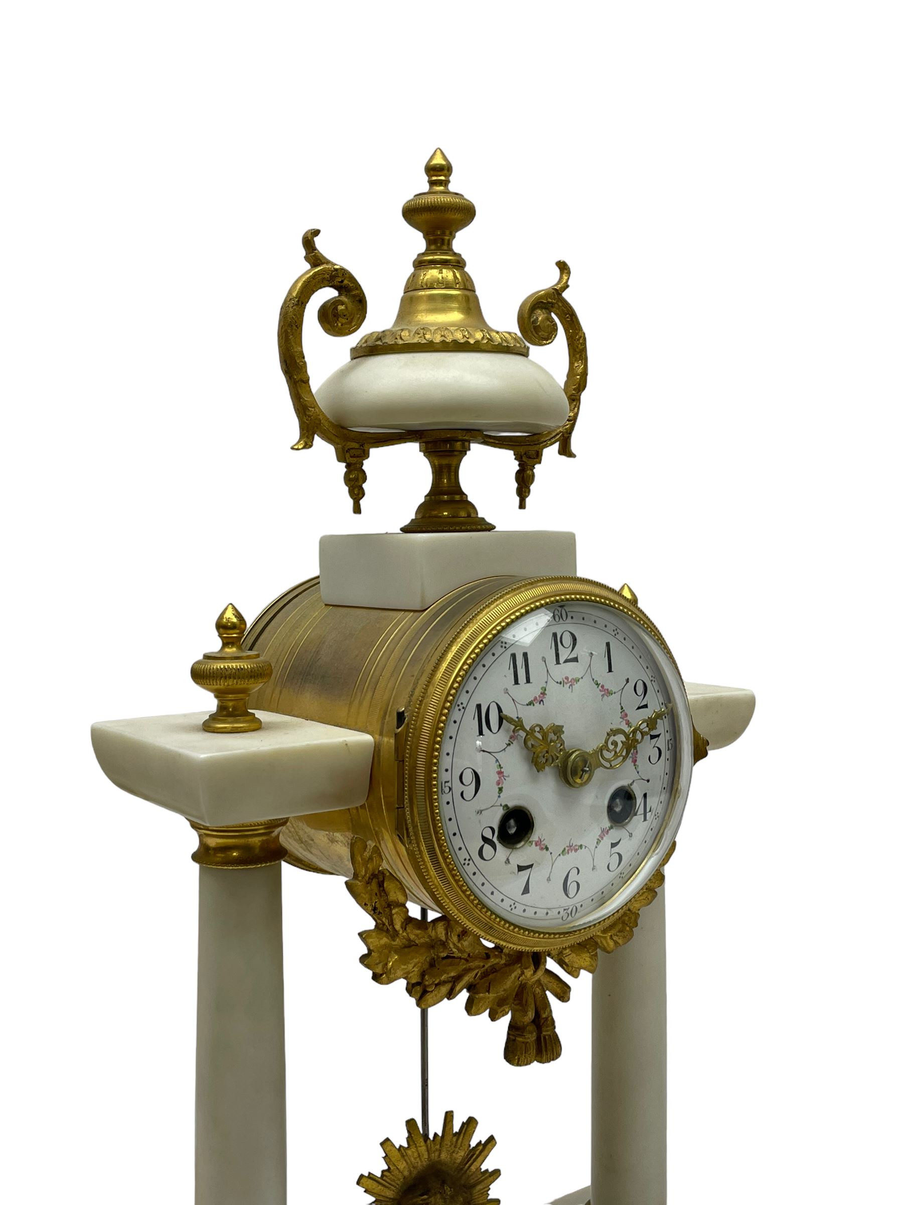 French - Late 19th century 8-day white marble and gilt metal portico clock garniture, the clock surmounted by an urn and raised on a pair of cylindrical pillars above a rectangular plinth base, white enamel dial with Arabic numerals and floral garlands, twin train countwheel striking movement, striking the hours and half hours on a bell, with a sunburst pendulum and gilt Louis XVI hands, garniture with matching twin light candelabra. With Key.   Candelabra H 30cm.