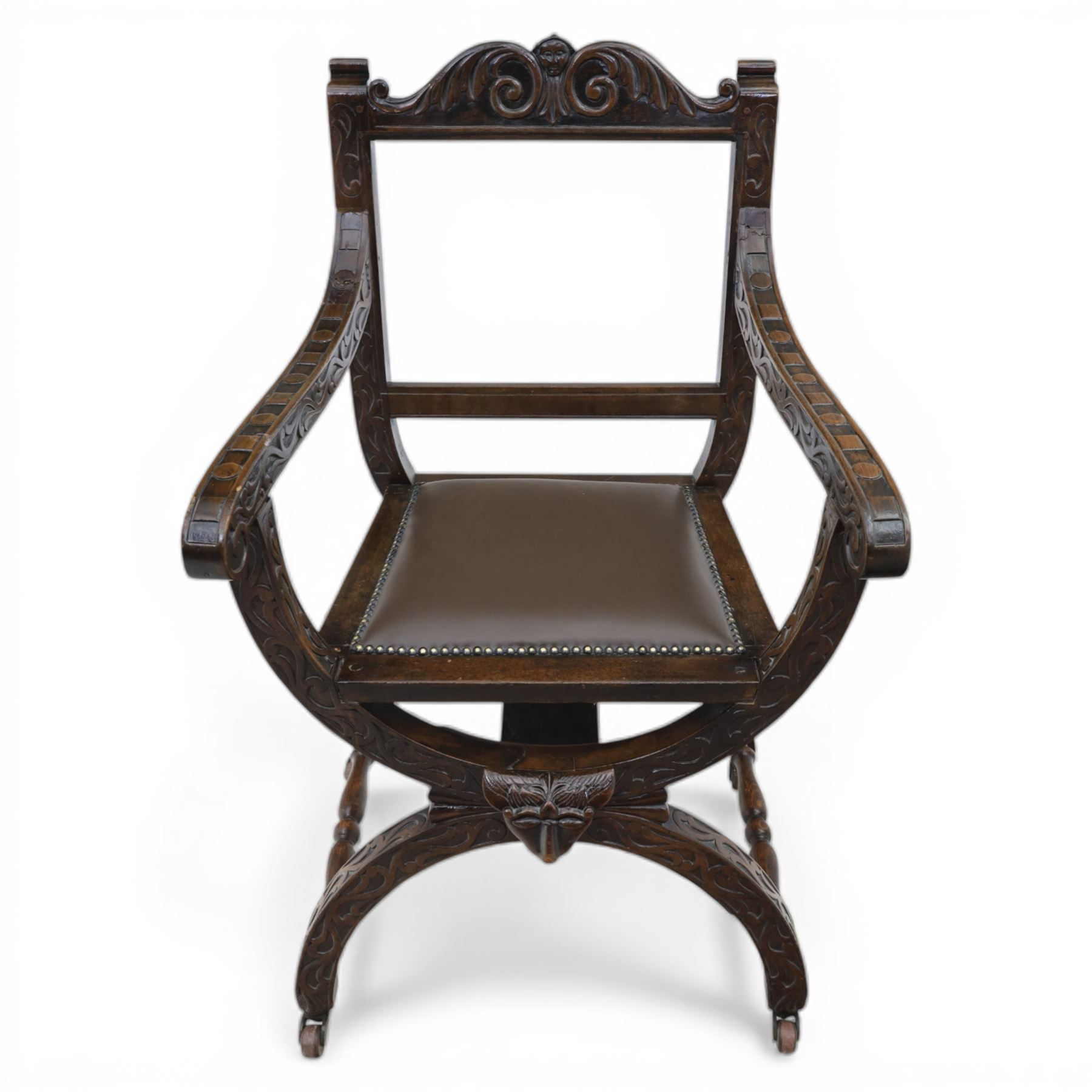 Late 20th century stained beech Savonarola chair, the carved crest rail and open arms above an upholstered leather seat, raised on an X framed base with carved foliate and mask decoration, joined by turned stretcher and ending in castors