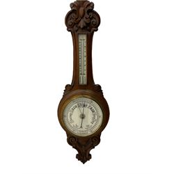 An English 1930’s solid oak carved hall barometer in a scroll shaped carved case with relief carving, aneroid movement, 8” white register measuring barometric air pressure from twenty-six to thirty-one point nine inches, weather predictions in black upper and lower case gothic script with a blue steel indicating hand and brass recording hand, brass dial bezel with flat bevelled glass, mercury thermometer enclosed in a glazed rectangular box displaying temperature in degrees Fahrenheit and Celsius on a white register. H87cm


