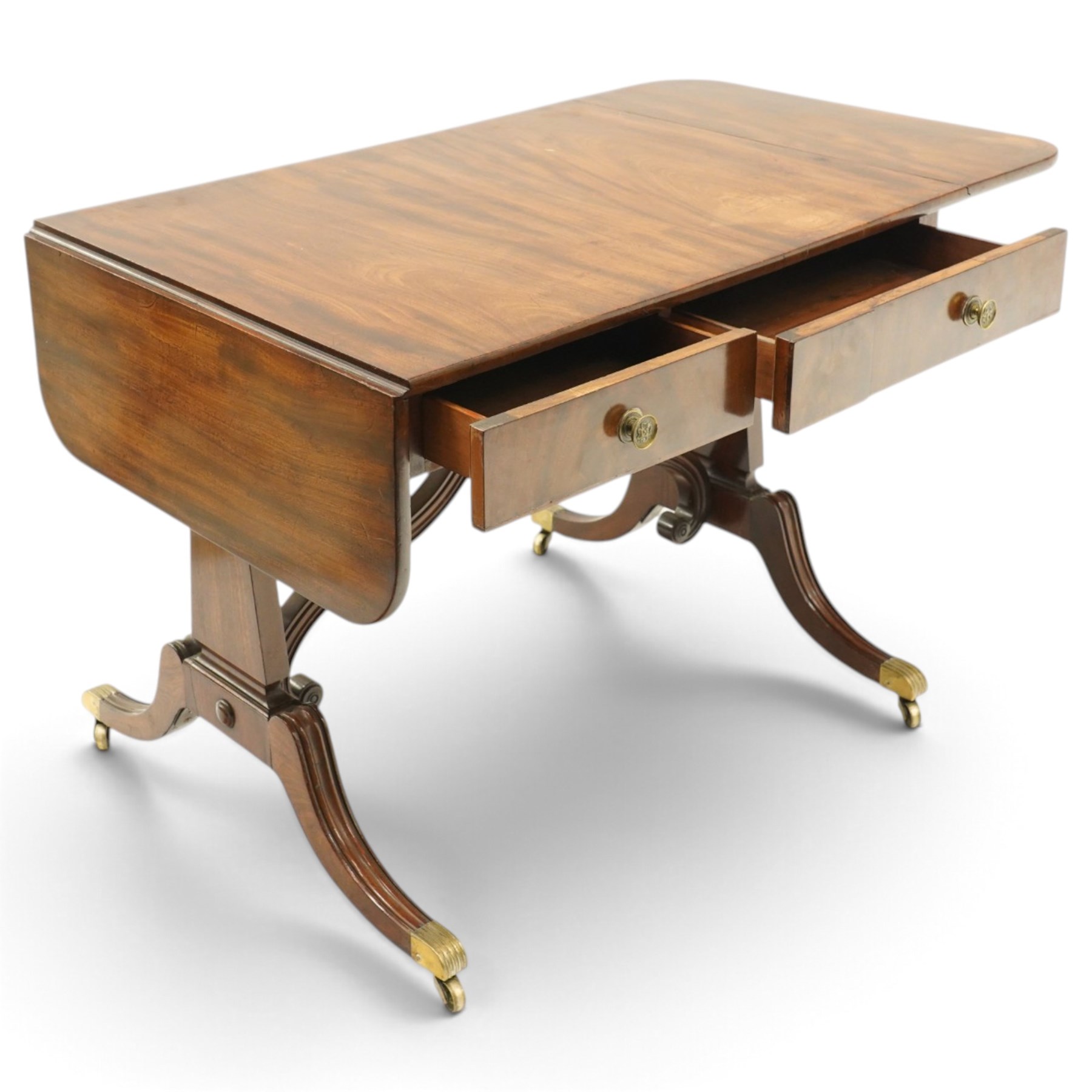 Early 20th century Regency Revival figured mahogany sofa table, rectangular drop-leaf top with rounded corners, fitted with two drawers to each side with brass flowerhead pull handles, raised on twin end supports with splayed reeded legs and brass cups and castors, united by a scrolling and reeded closed stretcher