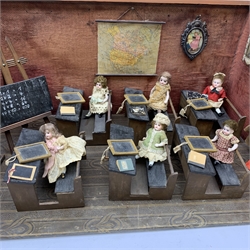 Late Victorian wooden school room setting with bisque head teacher and six German all bisque pupils each seated at a combined desk and chair with slate board, in a classroom furnished with chest of drawers, fire-surround, lectern, wall map, pictures, miniature books, school bell, chalk board on easel, wall clock etc, L70cm H28cm D28cm