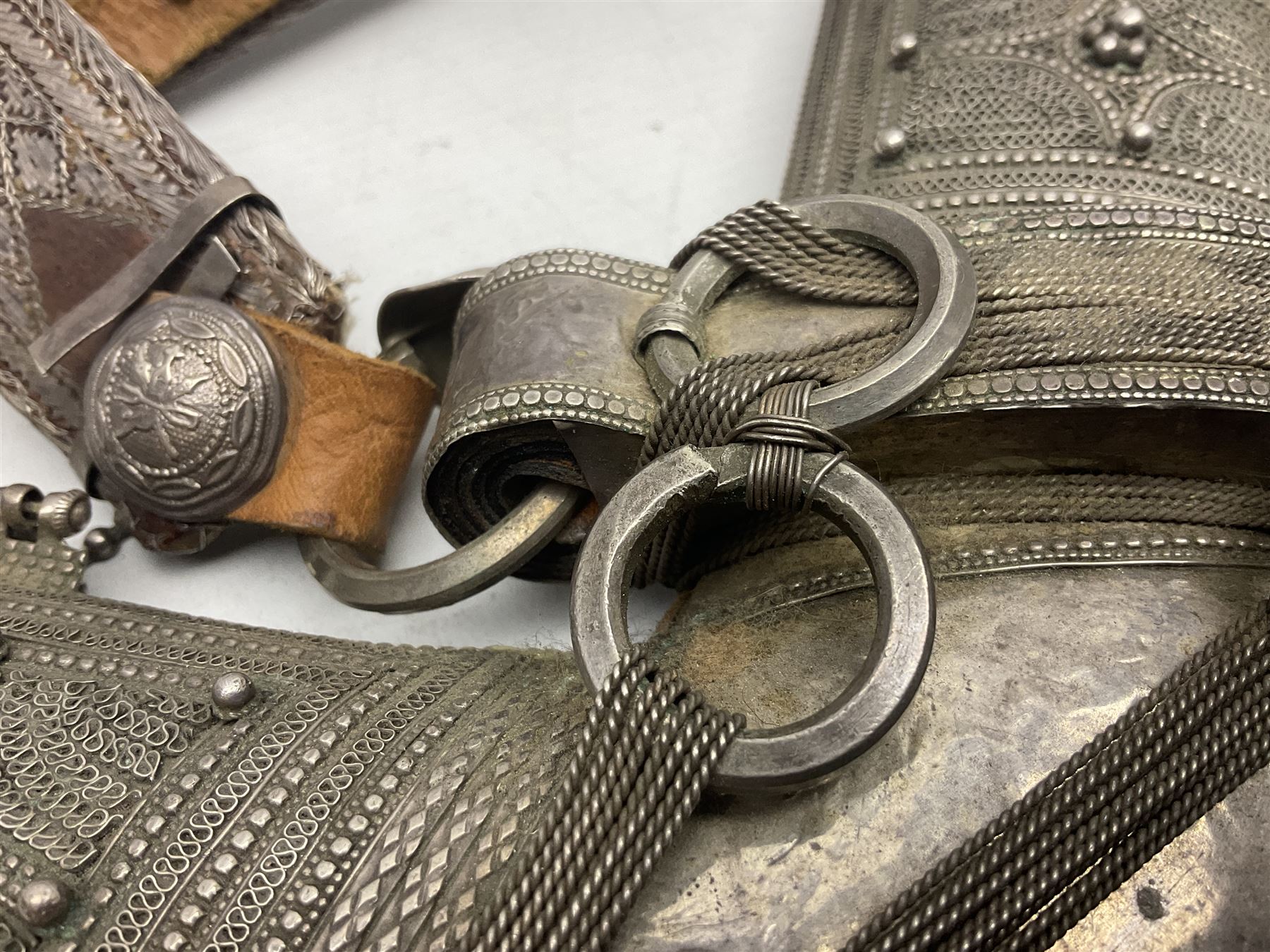 Omani Khanjar dagger, the 19cm curved blade with pronounced medial rib to either side, waisted hilt with dome shaped pommel and silver covered horn grip decorated with filigree bands, floral motifs and beadwork; in an intricately filigree decorated silver and leather scabbard with silver rings woven into the belt mount and fitted with original silver thread-work belt L34cm overall belt length 94cm