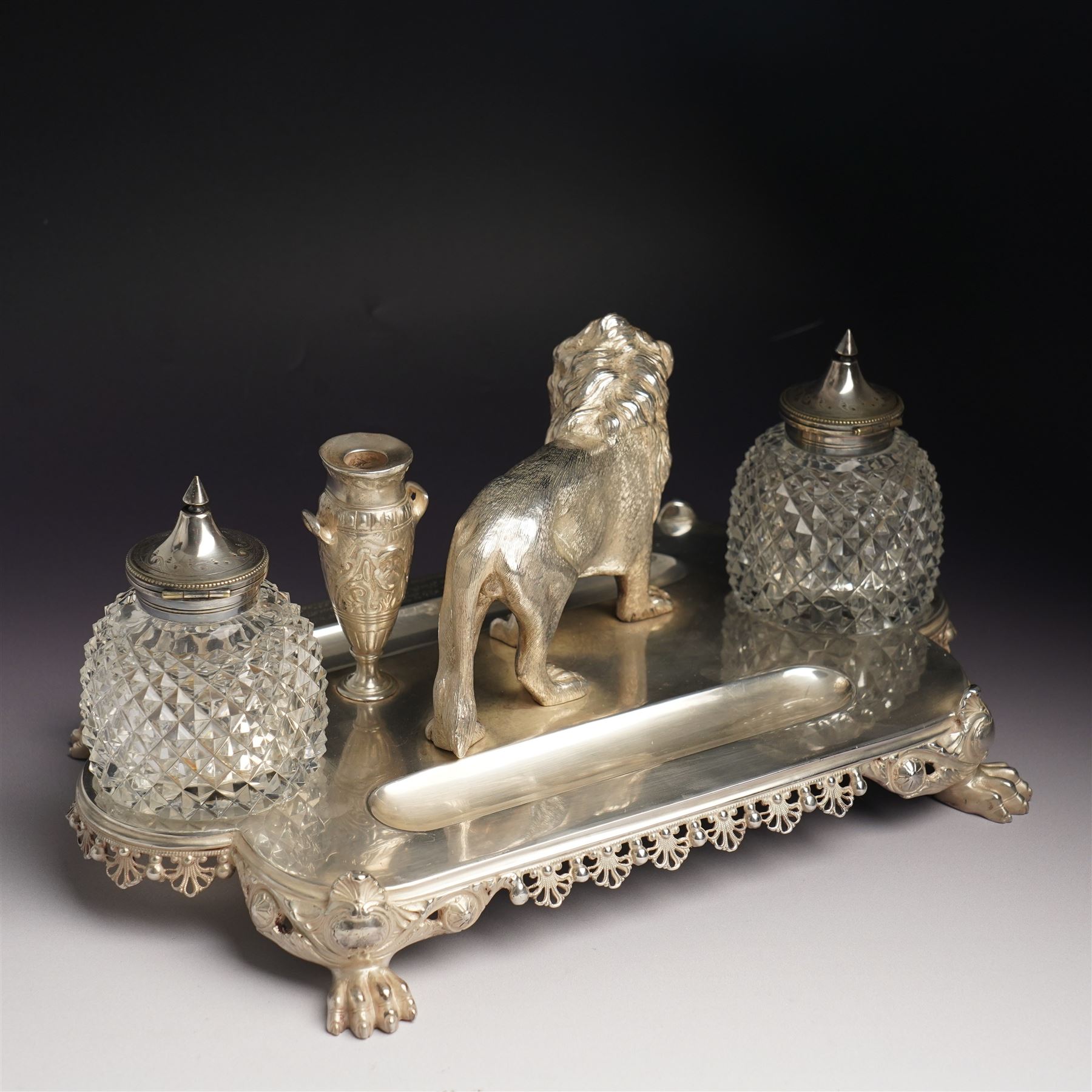 19th century Walker & Hall, silver plated inkstand, oblong and on four paw feet, the border decorated with pierced shell design, each side with a pen-rest, with two cut-glass bottles and central lion and urn finials, engraved to the front with an inscription, H15cm, L30cm