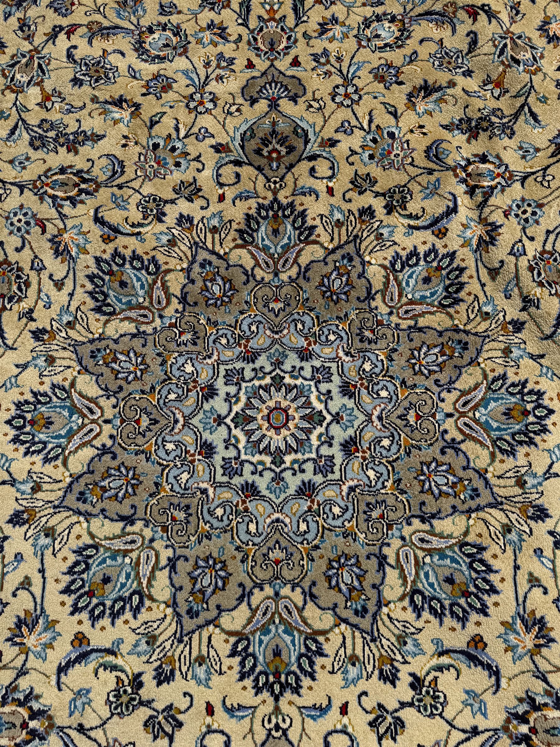 Central Persian Kashan pale khaki and ivory ground carpet, shaped central medallion enclosing small stylised motifs, the field decorated profusely with trailing foliate branches and plant motifs, guarded border with repeating floral design