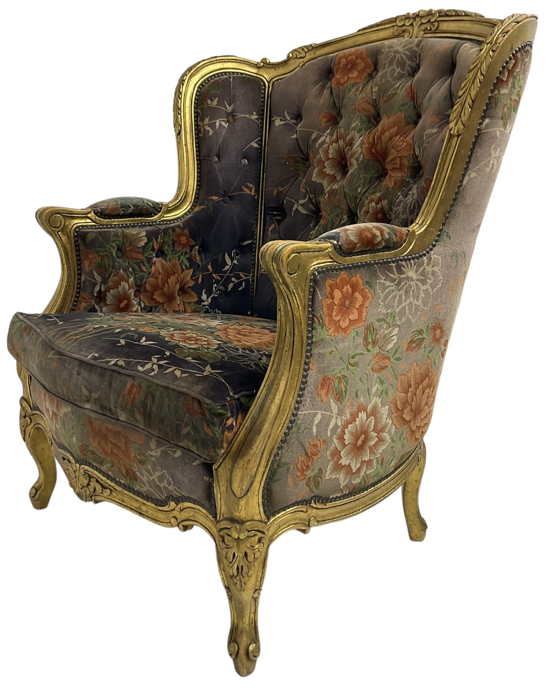 Pair of Louis XVI design gilt framed wingback armchairs, cresting rail carved and moulded with foliate decoration and C-scrolls, upholstered in buttoned floral patterned mauve velvet with loose seat cushion, scallop carved apron over cabriole supports